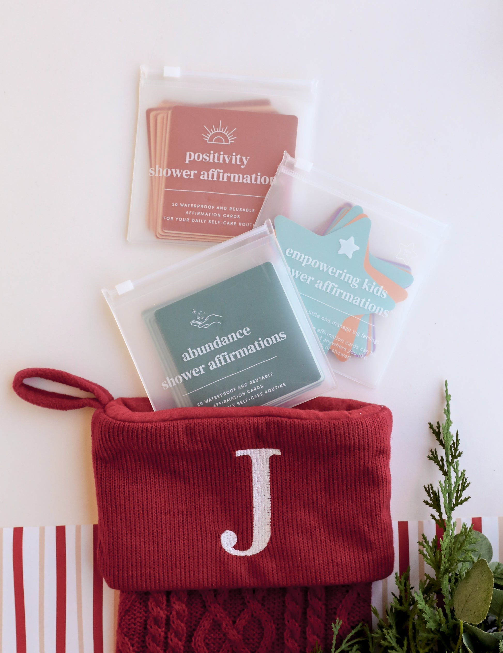 A red knit stocking with a white J holds three packs of affirmation cards labeled positivity, abundance, and empowering kids, next to holiday greenery and striped wrapping paper.