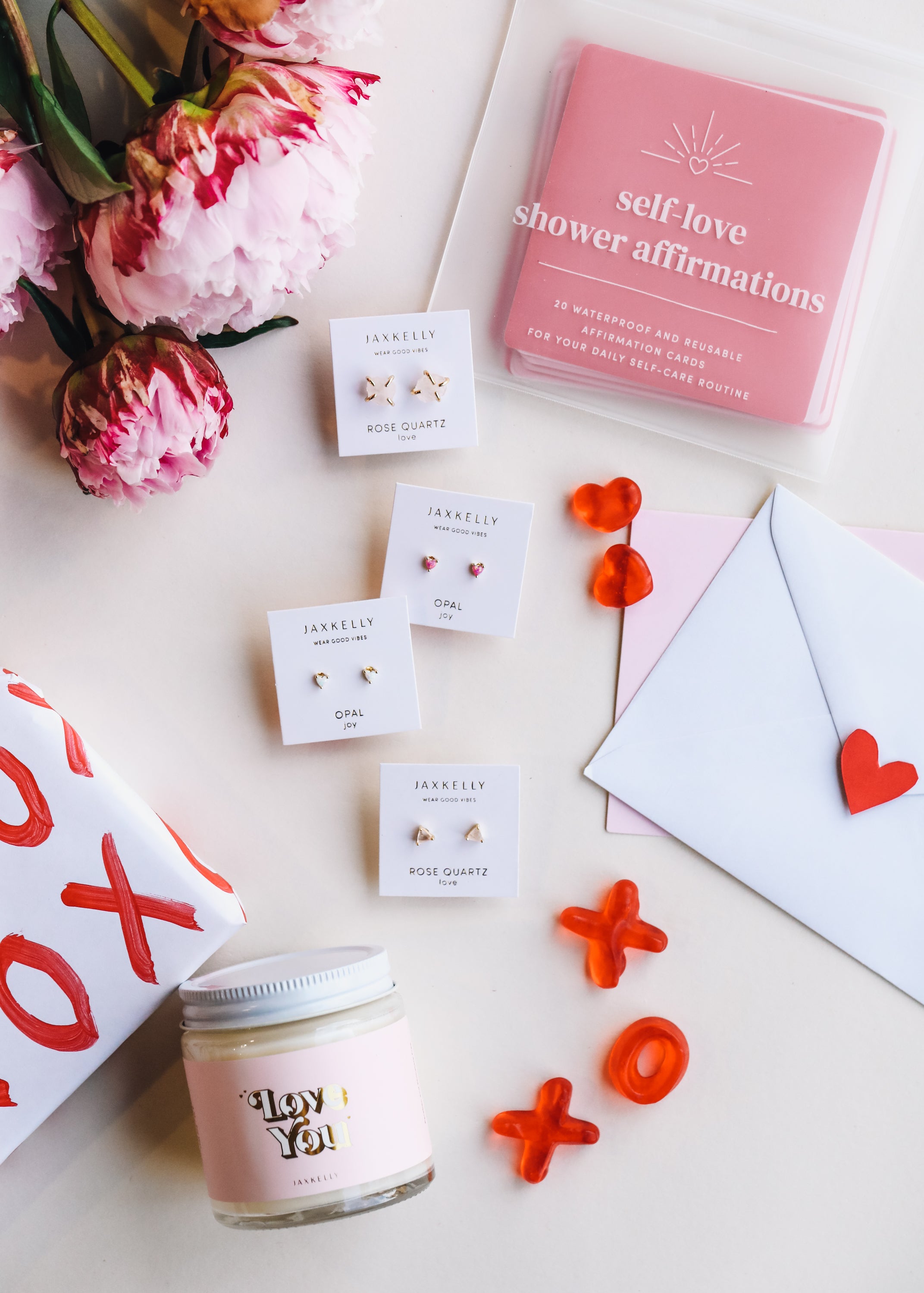 Flat lay of self-care gifts: pink peonies, affirmation cards, love-themed jewelry, jar candle labeled Love You, red gummy candies, and an envelope with a heart sticker on a white surface.