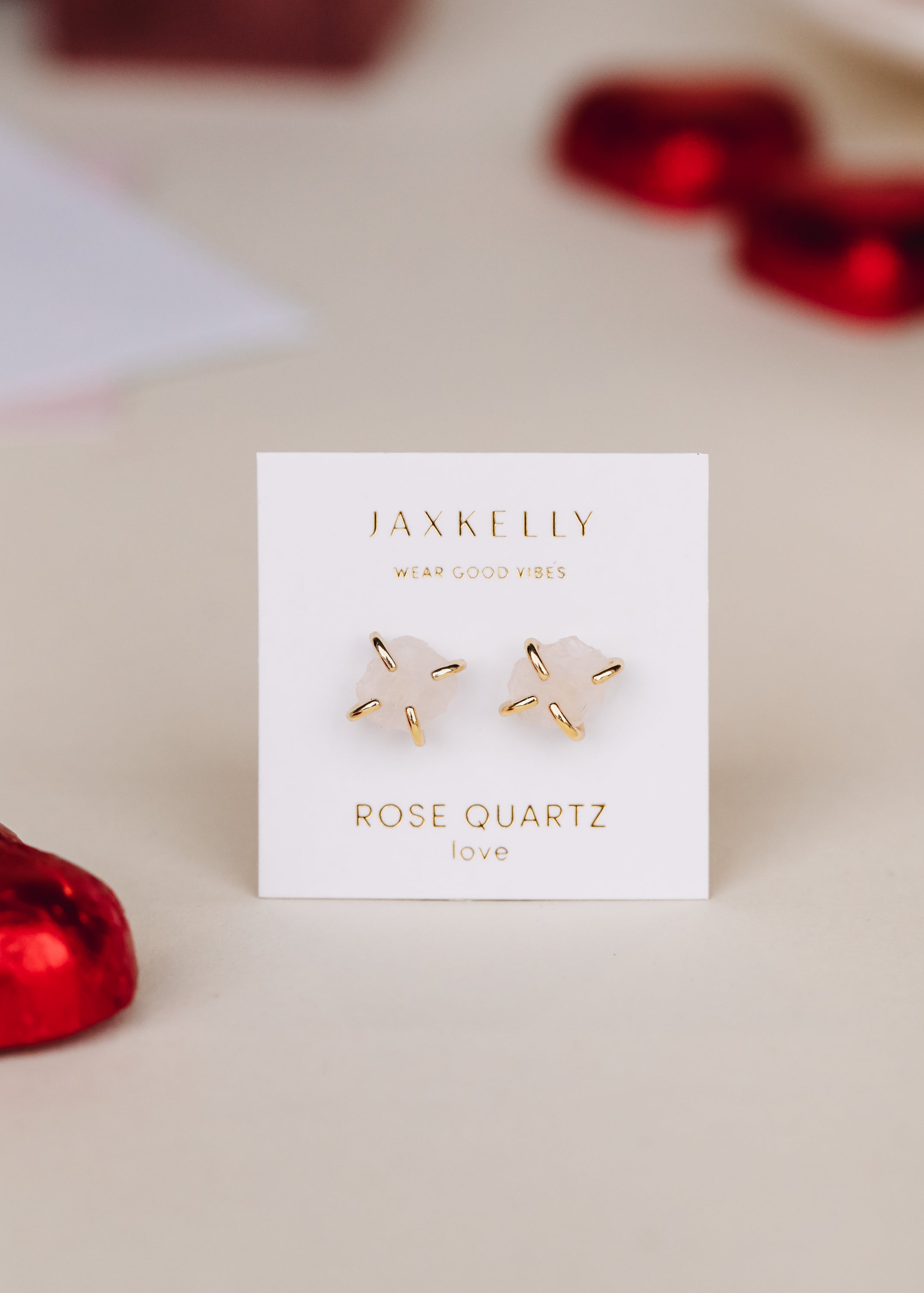 A pair of gold and rose quartz earrings displayed on a white JaxKelly card, surrounded by red heart-shaped objects on a beige surface.