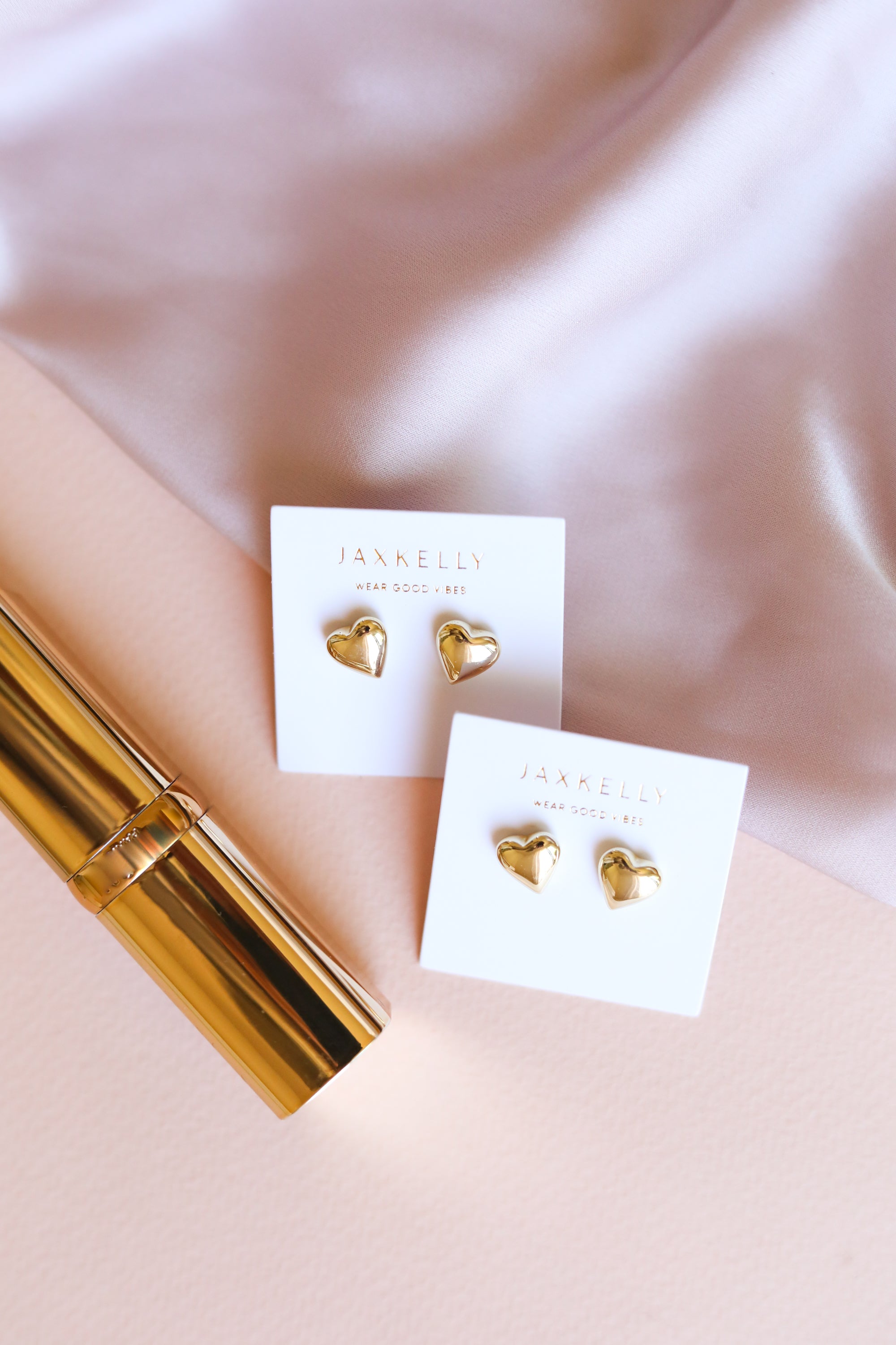 Two pairs of gold heart-shaped earrings on white JaxKelly cards are displayed on a soft pink surface with a gold tube and a silky light pink fabric nearby.