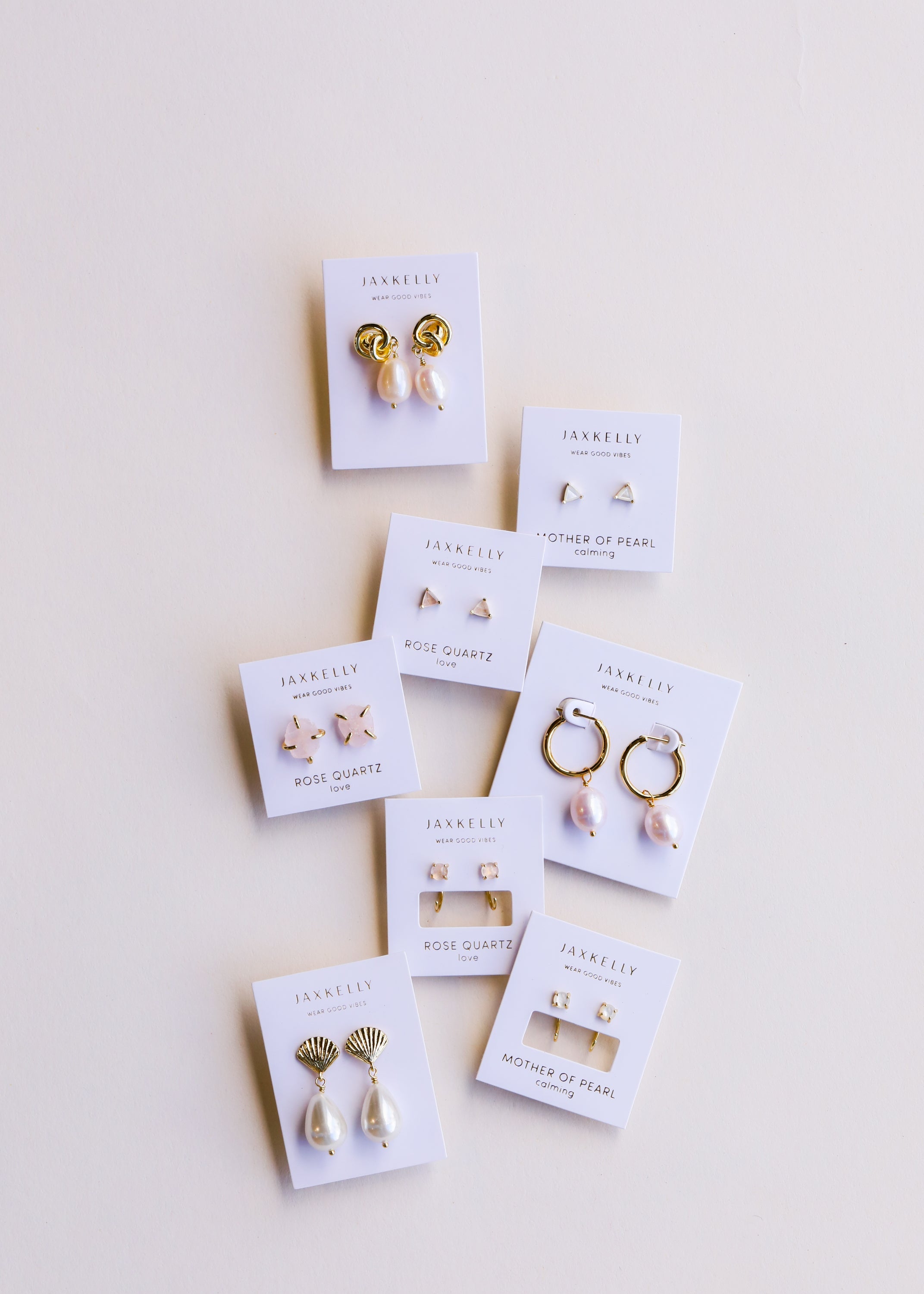 Nine pairs of elegant earrings, each on a white JaxKelly card, are arranged on a light background. The earrings feature gold, pearls, and gemstones in various styles.
