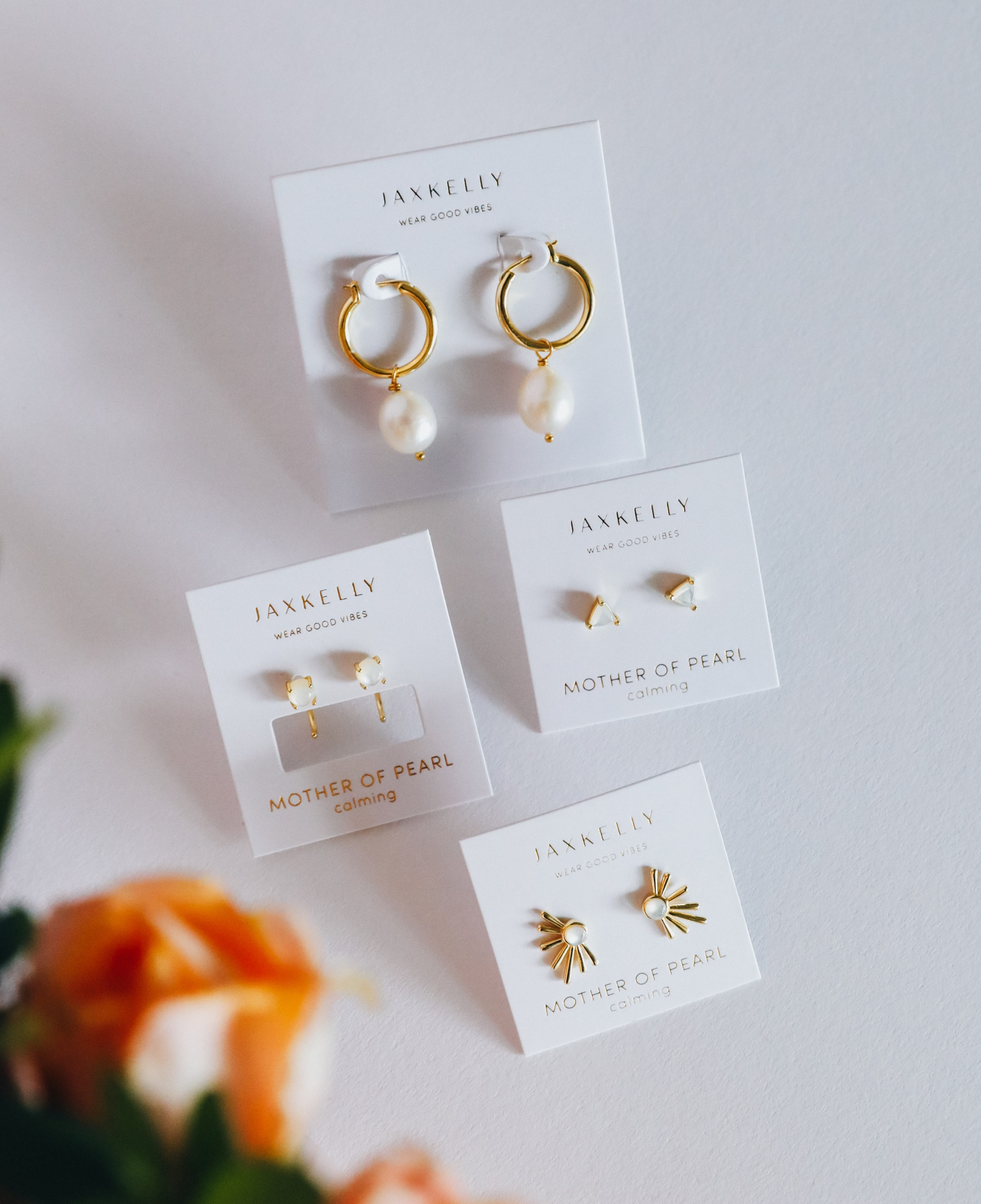 A flat lay of four pairs of gold and mother of pearl earrings by JaxKelly on white cards, arranged on a white surface with orange flowers in the corner.