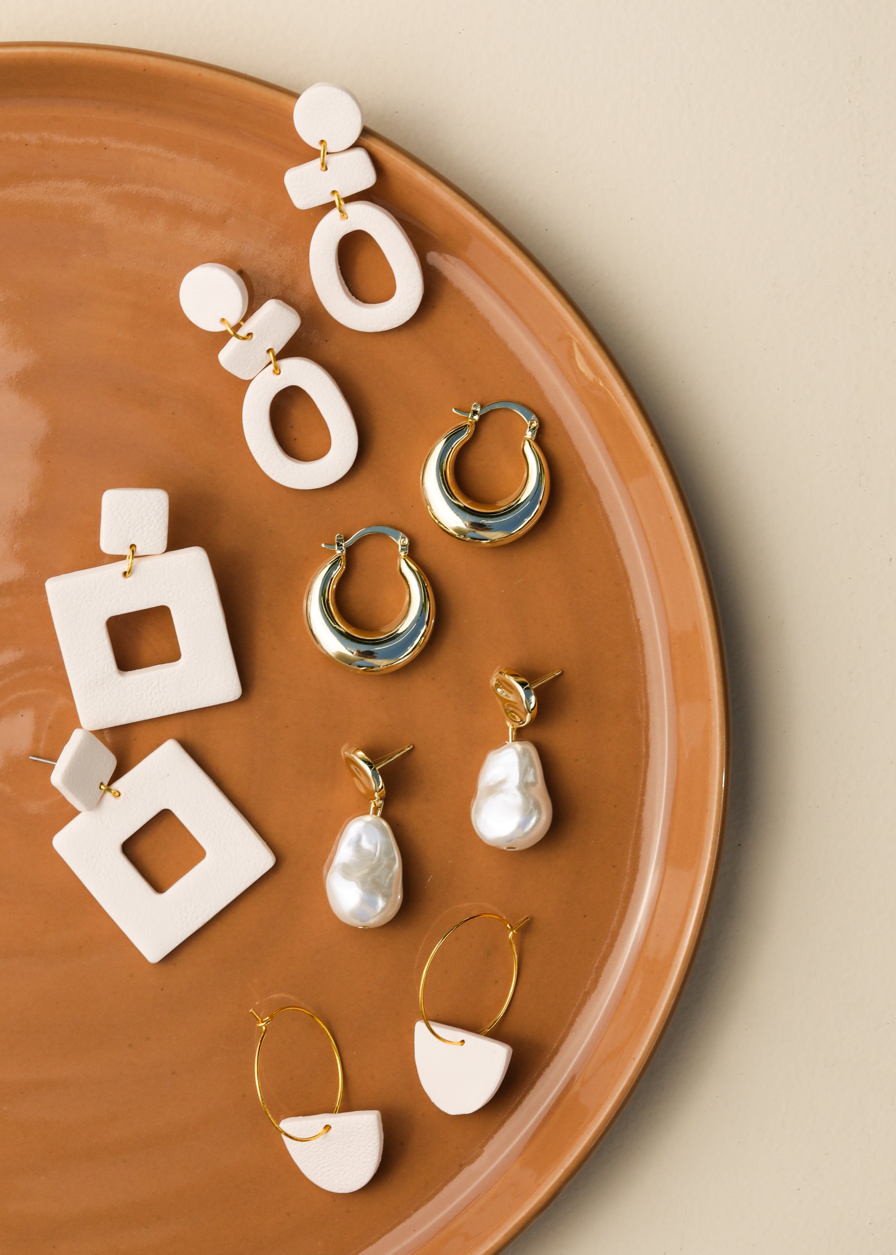 A terracotta plate displays five pairs of earrings: geometric white shapes, gold hoops, gold hoops with pearls, and white and gold semicircle designs, all arranged neatly on a beige background.