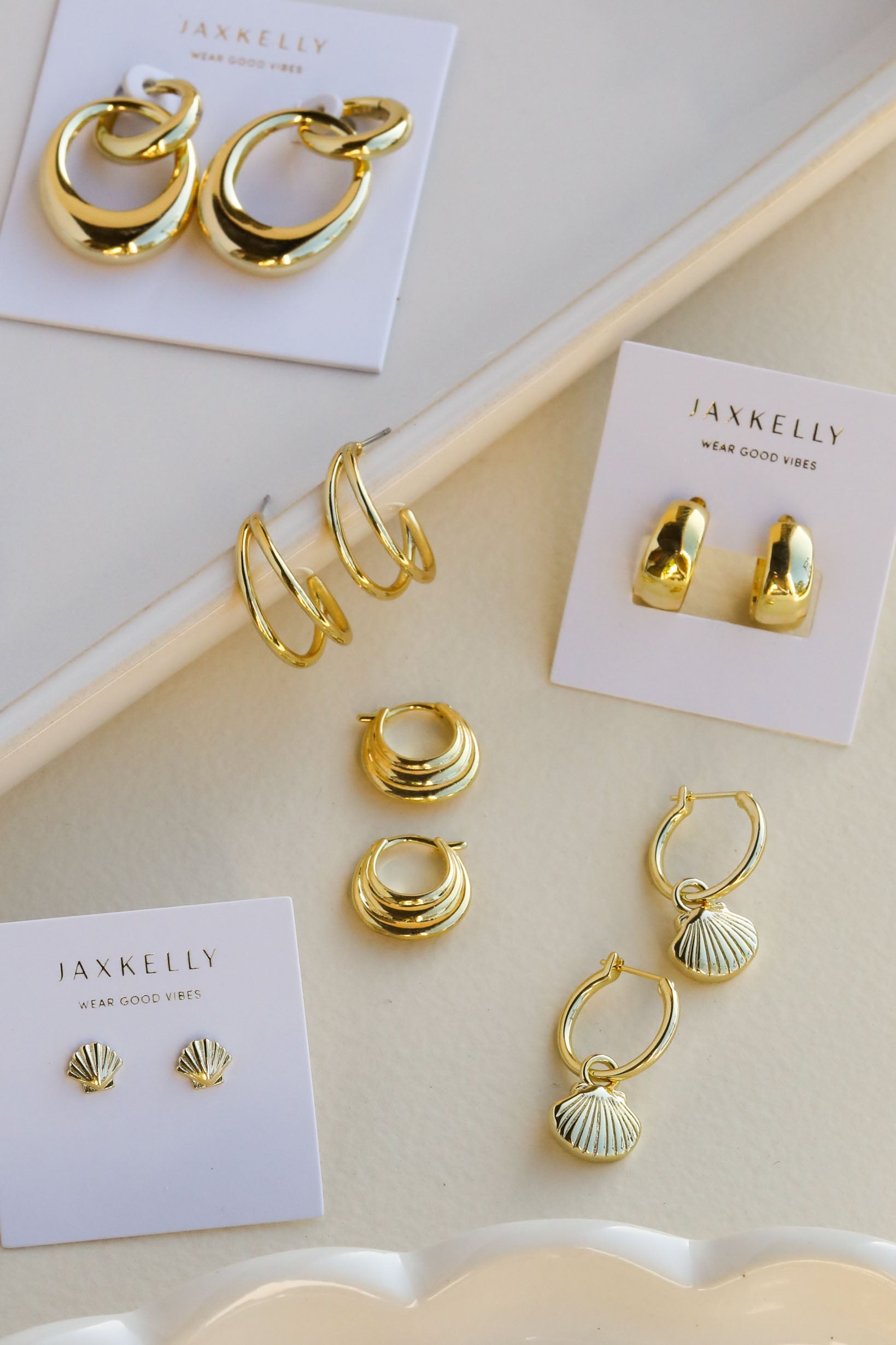 A display of six pairs of gold earrings, including hoops, studs, and shell designs, arranged neatly on a cream surface with three pairs on branded JaxKelly cards.