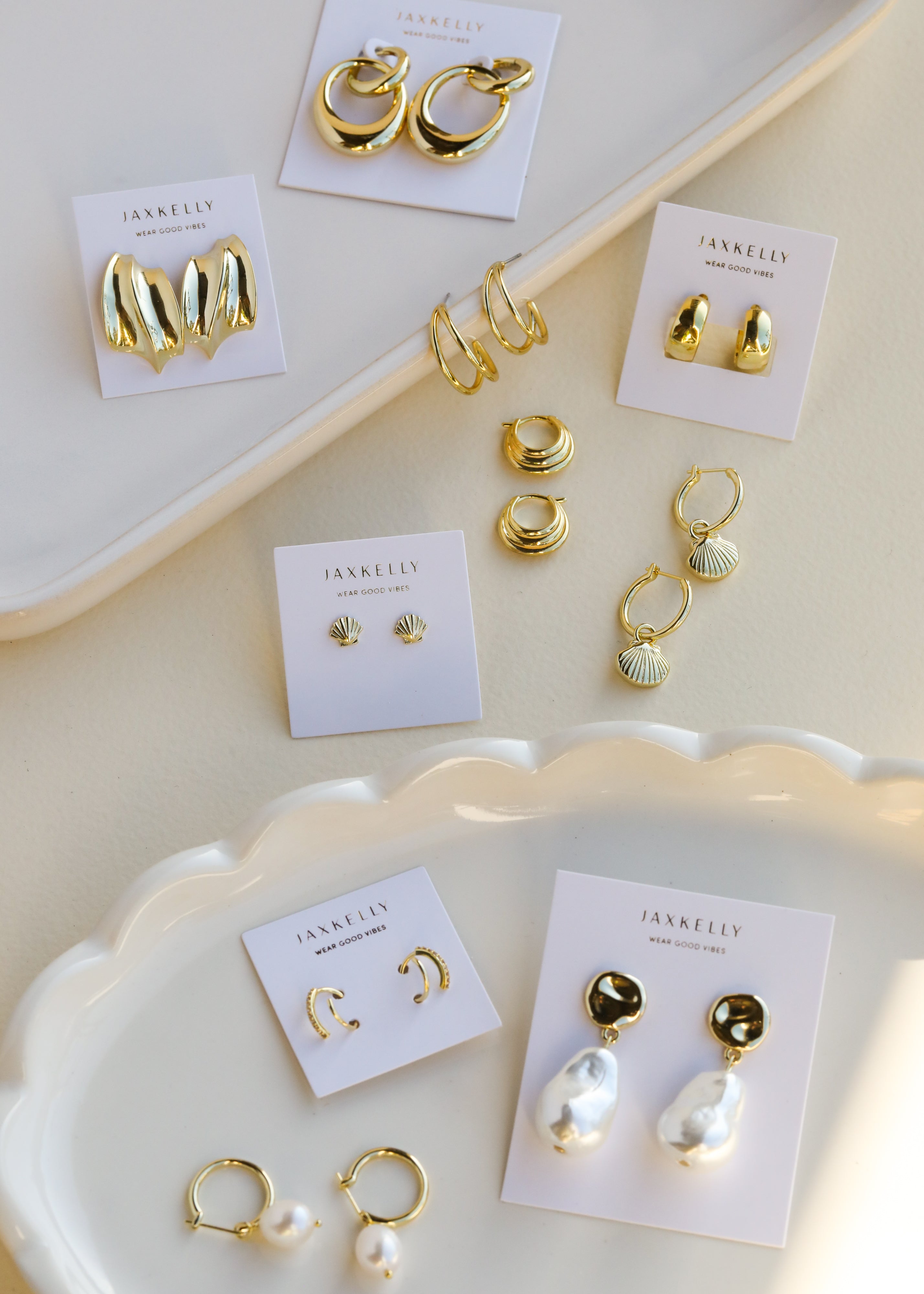 Assorted gold and pearl earrings on display cards are arranged on white ceramic trays, including hoops, shells, studs, and drop styles, all labeled with the brand name JaxKelly.