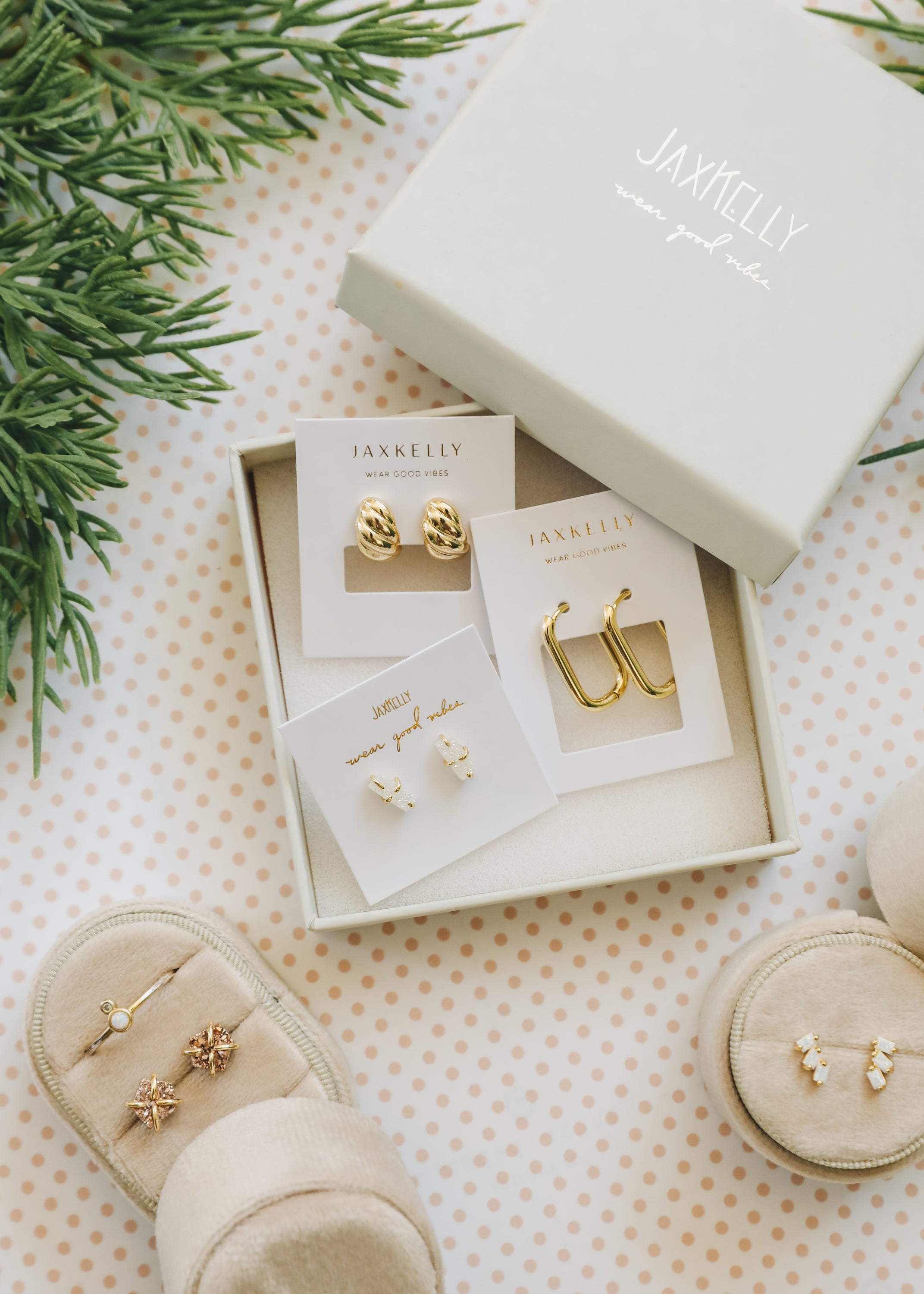 A white JaxKelly jewelry box holds three pairs of gold earrings on display cards. Nearby are beige earring holders with more earrings, all on a polka-dot surface with green pine branches in the corner.