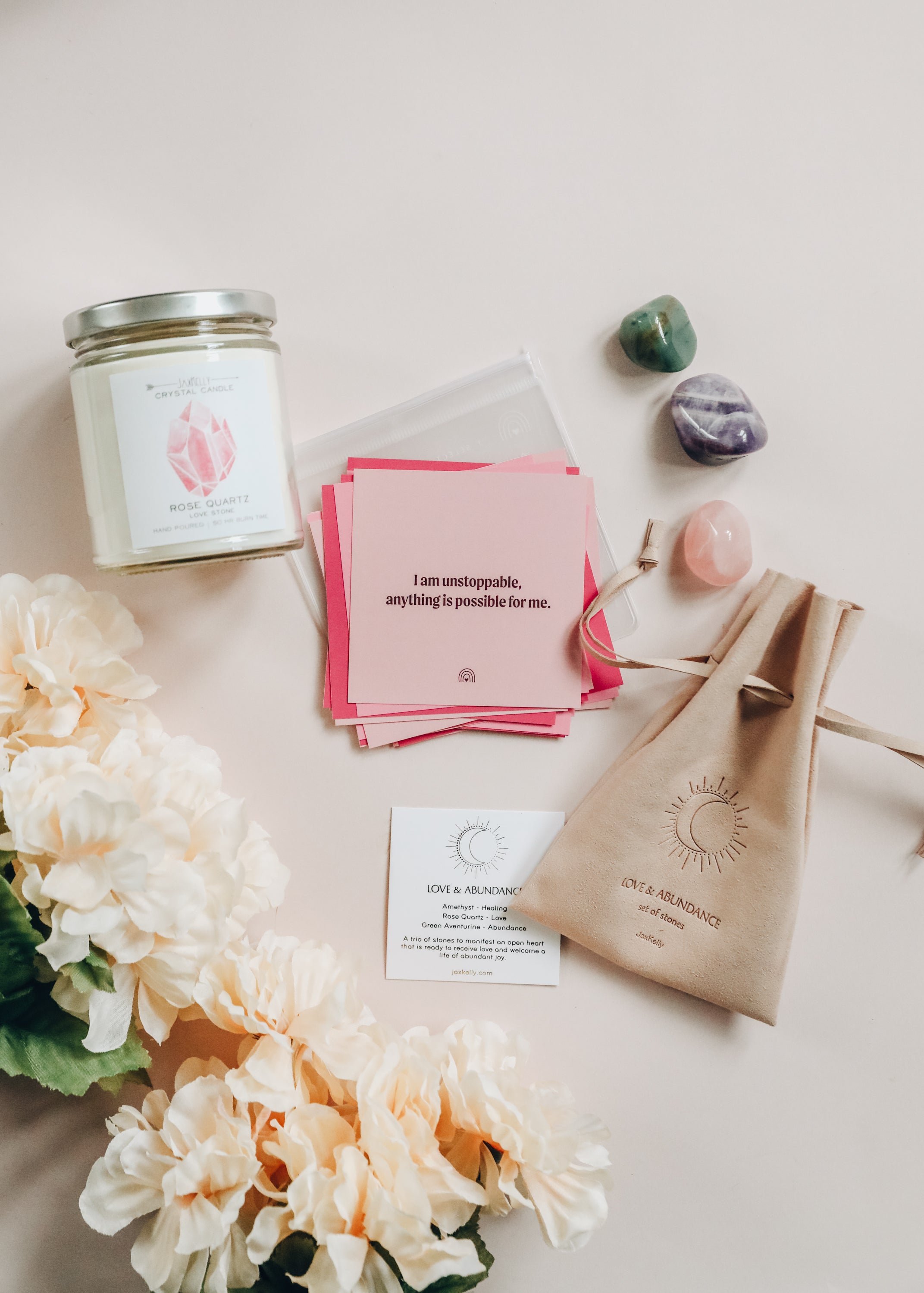 A flat lay of a candle, affirmation cards, three crystals, a beige pouch, a card, and pale peach flowers arranged on a light surface. The top card reads, I am unstoppable, anything is possible for me.