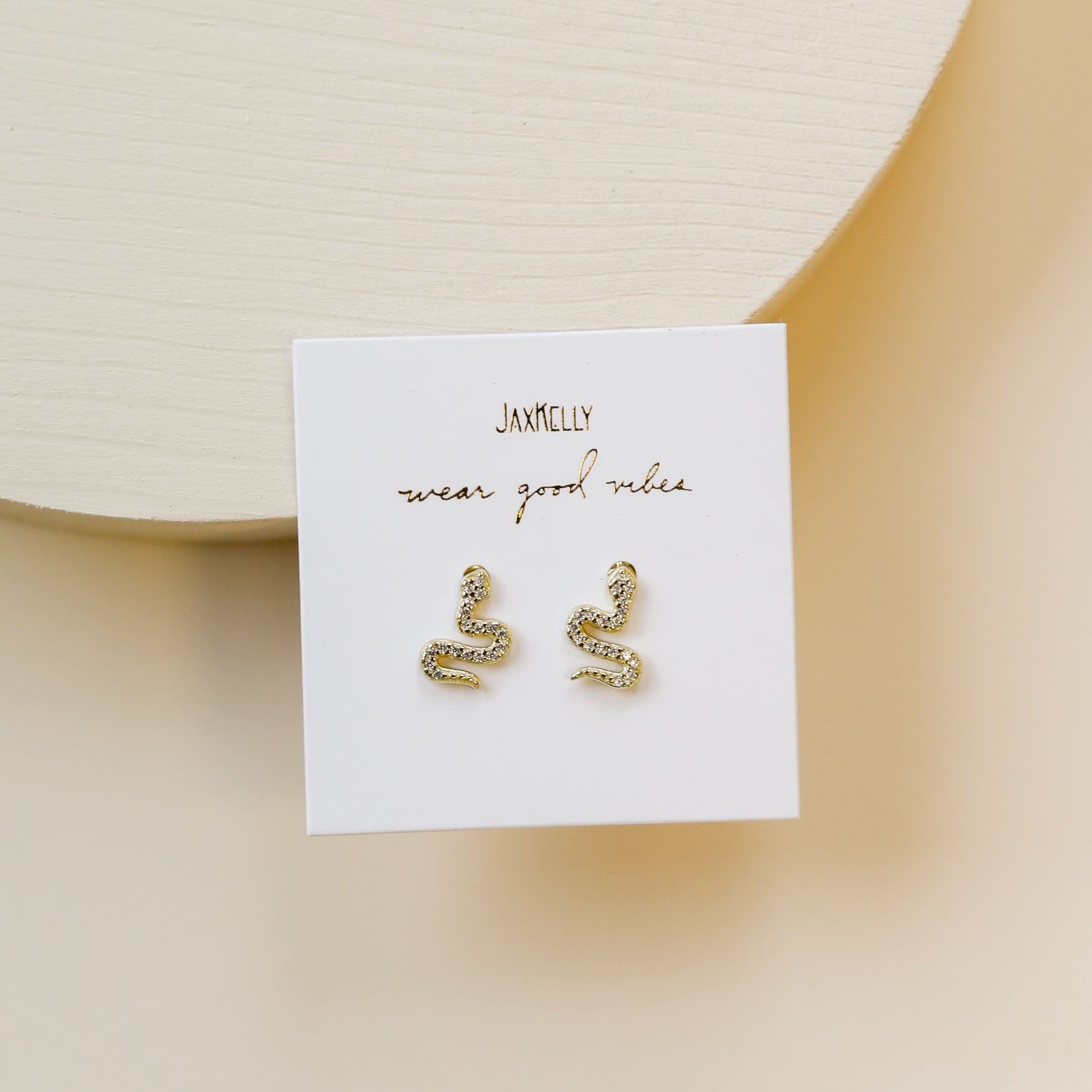 Gold snake-shaped earrings with sparkling details displayed on a white card labeled JaxKelly wear good vibes, set against a light beige textured background.