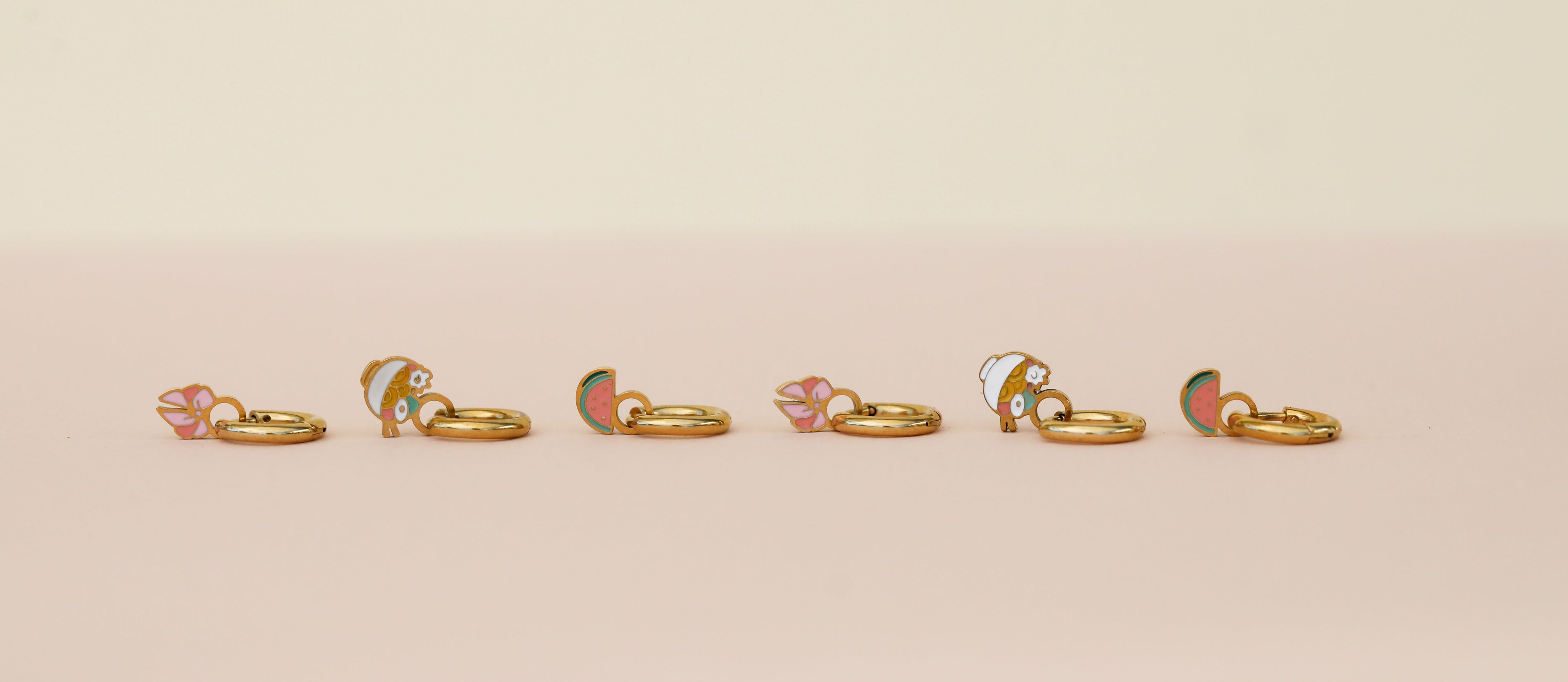 Six small gold hoop earrings with colorful enamel charms, including flowers and fruit shapes, are arranged in a row on a pink and cream background.