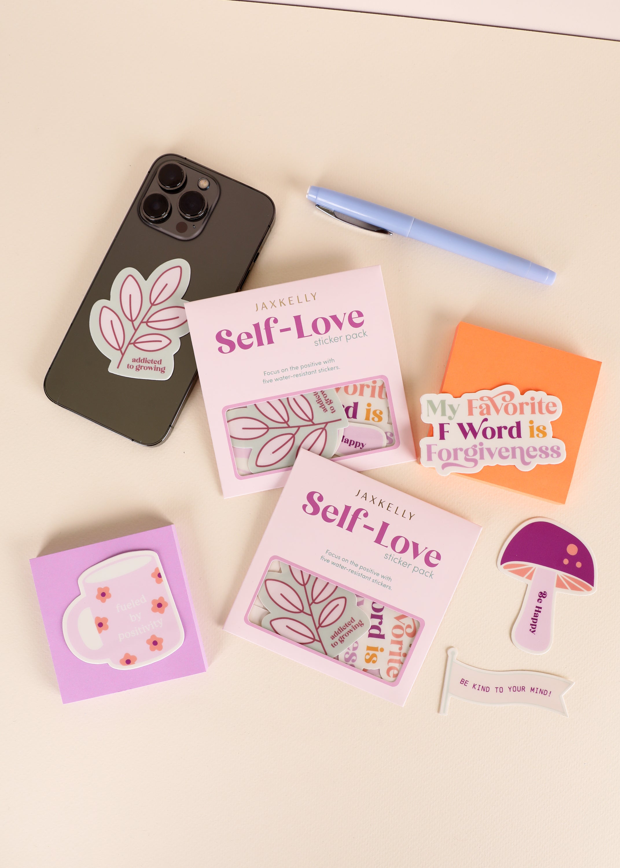 Flat lay of a phone with a leaf sticker, a blue pen, orange sticky note with My Favorite F Word is Forgiveness, two self-love sticker packs, a mug sticker, and a purple mushroom sticker.