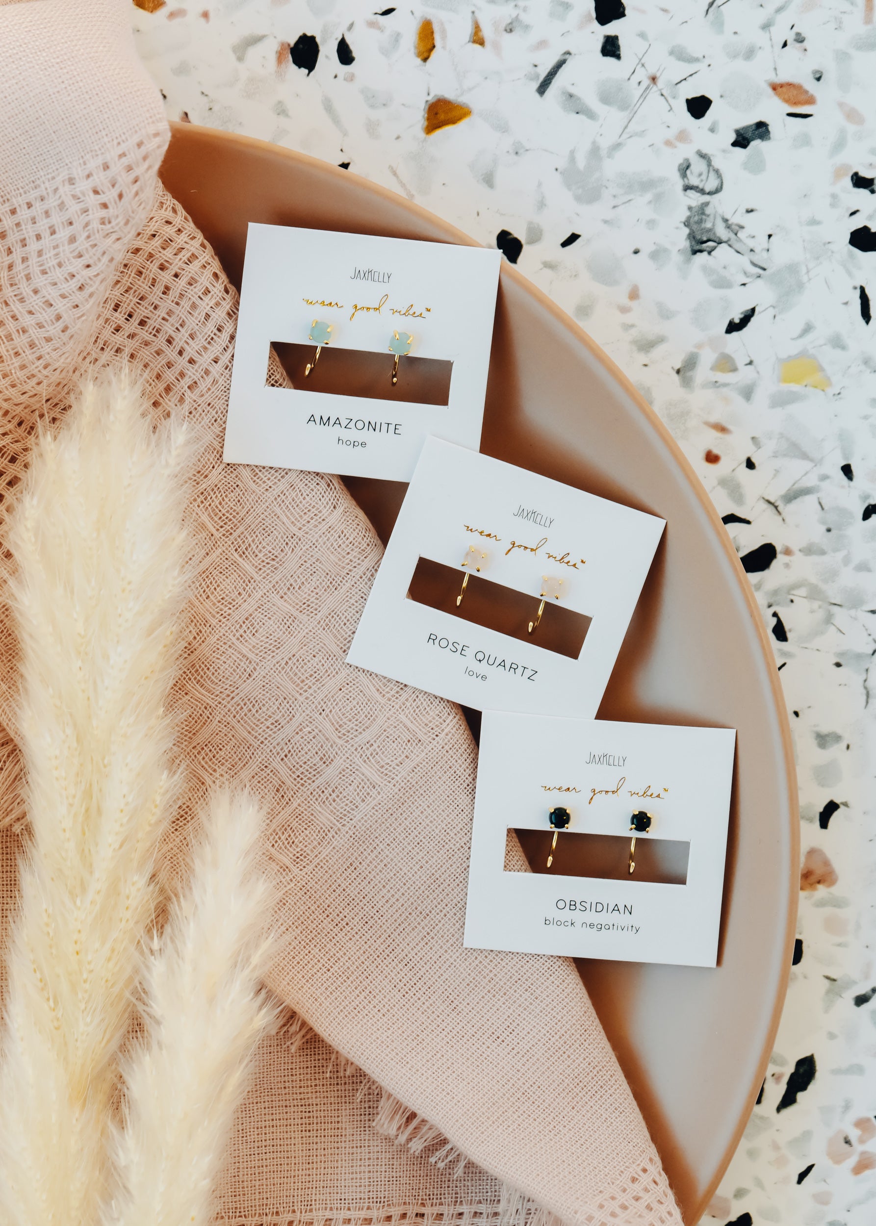 Three pairs of gemstone earrings—amazonite, rose quartz, and obsidian—are displayed on earring cards atop a pink textured fabric and a beige plate, with pampas grass and a terrazzo background.