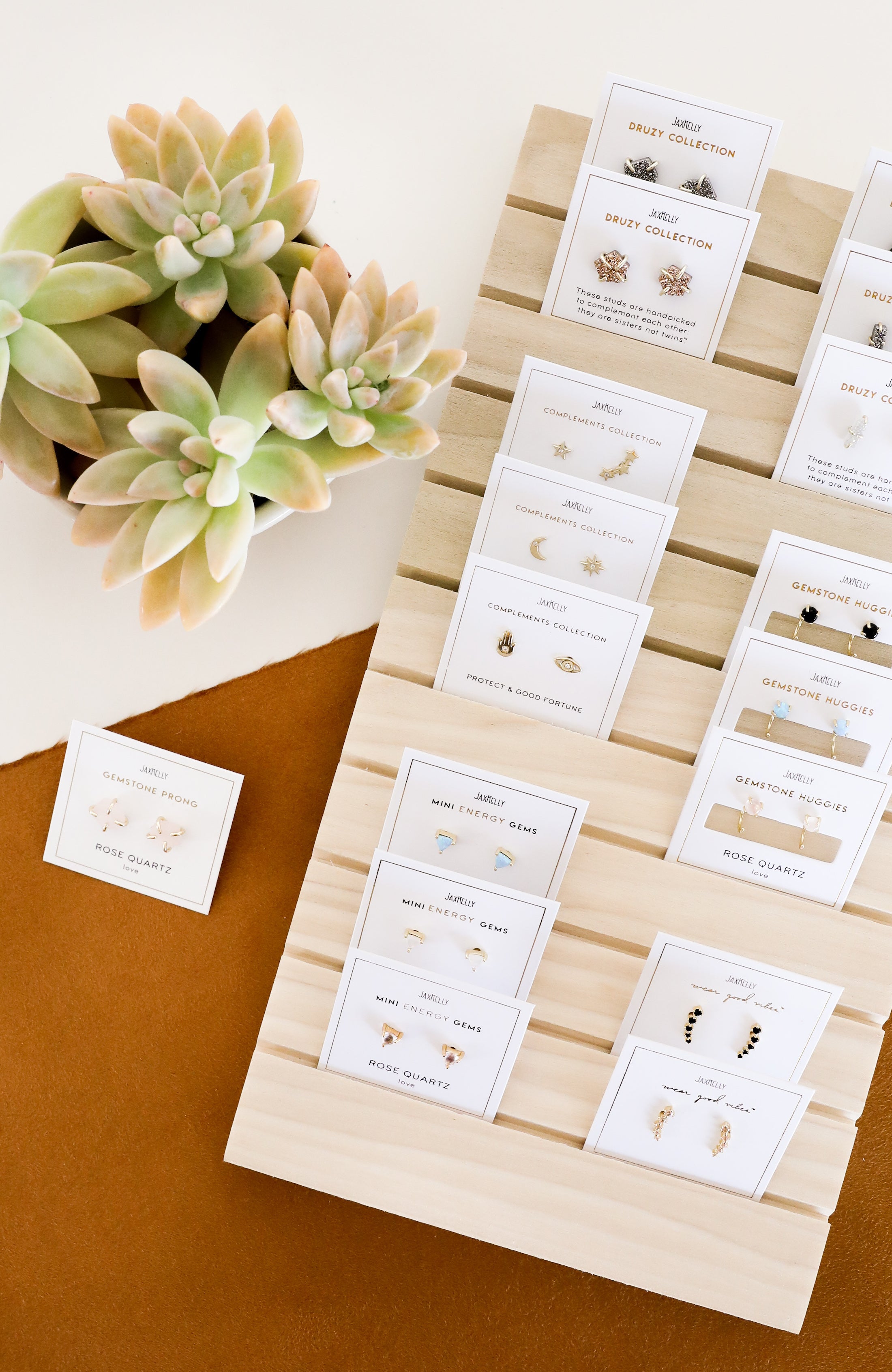A wooden display holds multiple pairs of earrings on white cards, next to a potted succulent plant. The display is set on a brown and white surface.