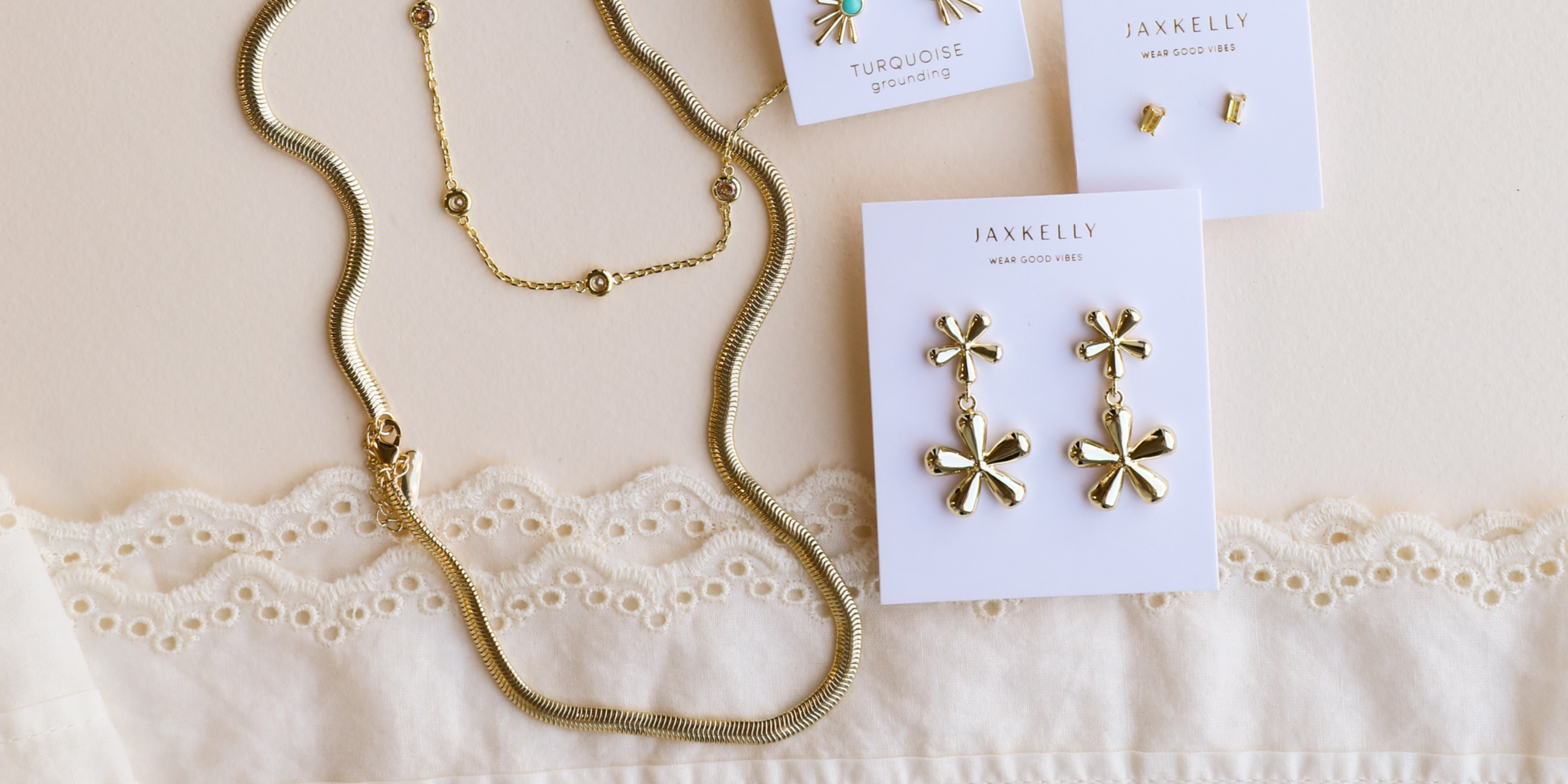 Gold necklaces and earrings displayed on a lace-trimmed fabric, with branded cards holding flower-shaped earrings, small studs, and turquoise jewelry on a cream background.