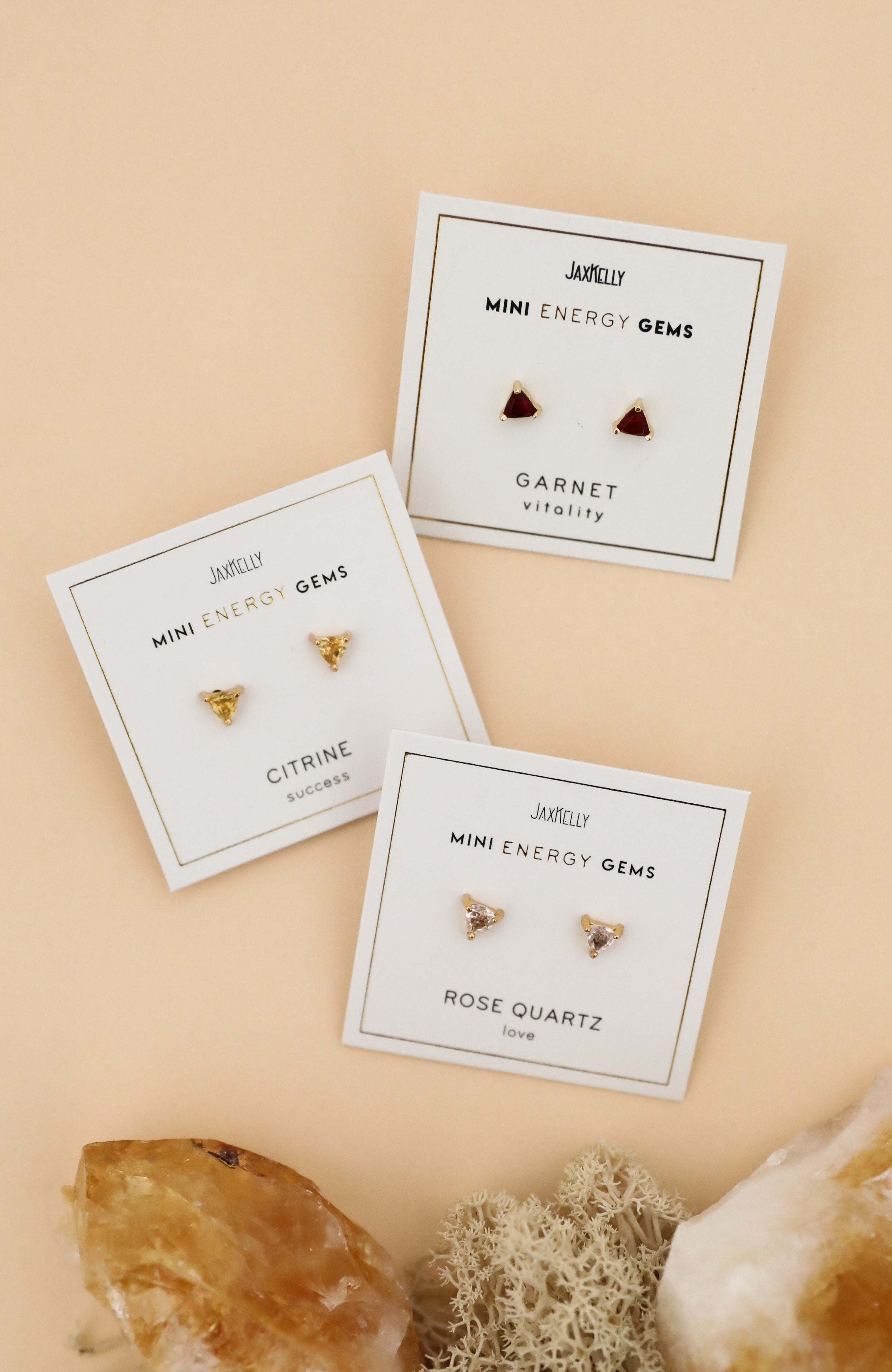Three pairs of stud earrings on cards labeled Mini Energy Gems: triangular citrine for success, heart-shaped garnet for vitality, and heart-shaped rose quartz for love, displayed with crystals on a beige background.