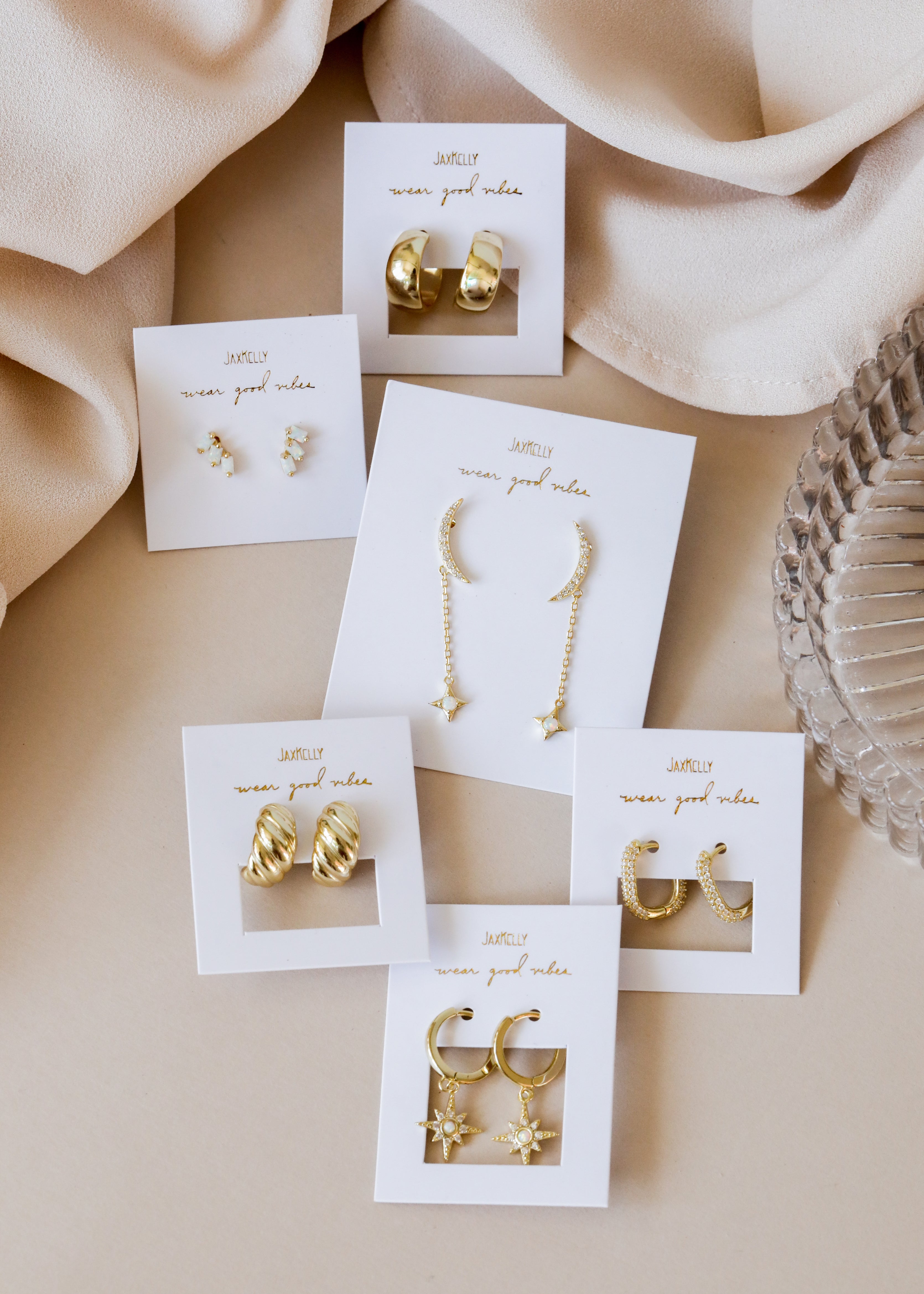 Six pairs of gold earrings, each on a white jewelry card labeled J&KELLY wear good vibes. Styles include hoops, studs, dangling stars, and crescent moons, arranged on a beige fabric background.