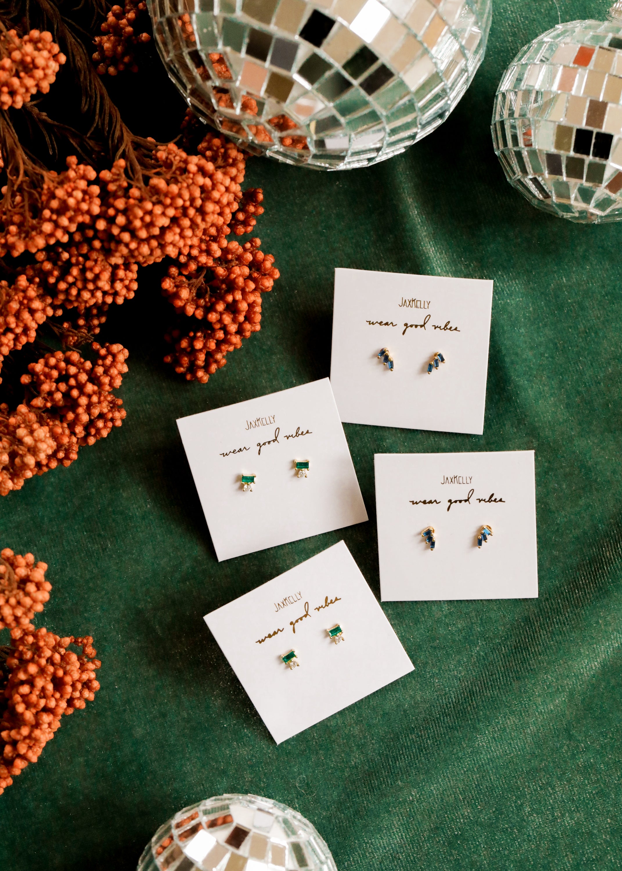 Four pairs of stud earrings on Wear Good Vibes cards are arranged on a green surface, surrounded by orange berries and mirrored disco balls.