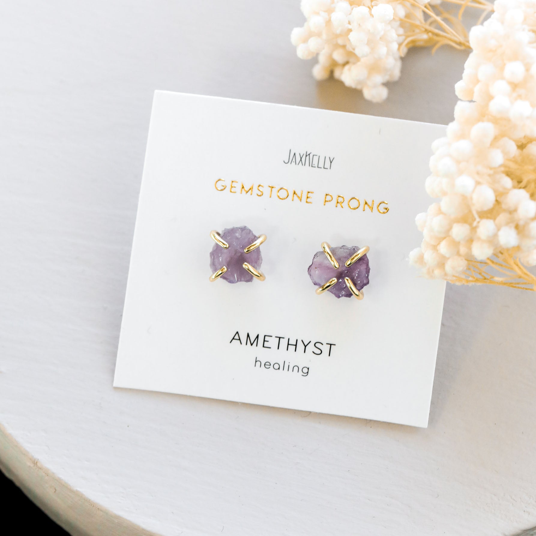 A pair of amethyst gemstone prong earrings with gold accents on a white card labeled Gemstone Prong Amethyst Healing, placed on a light surface near white dried flowers.