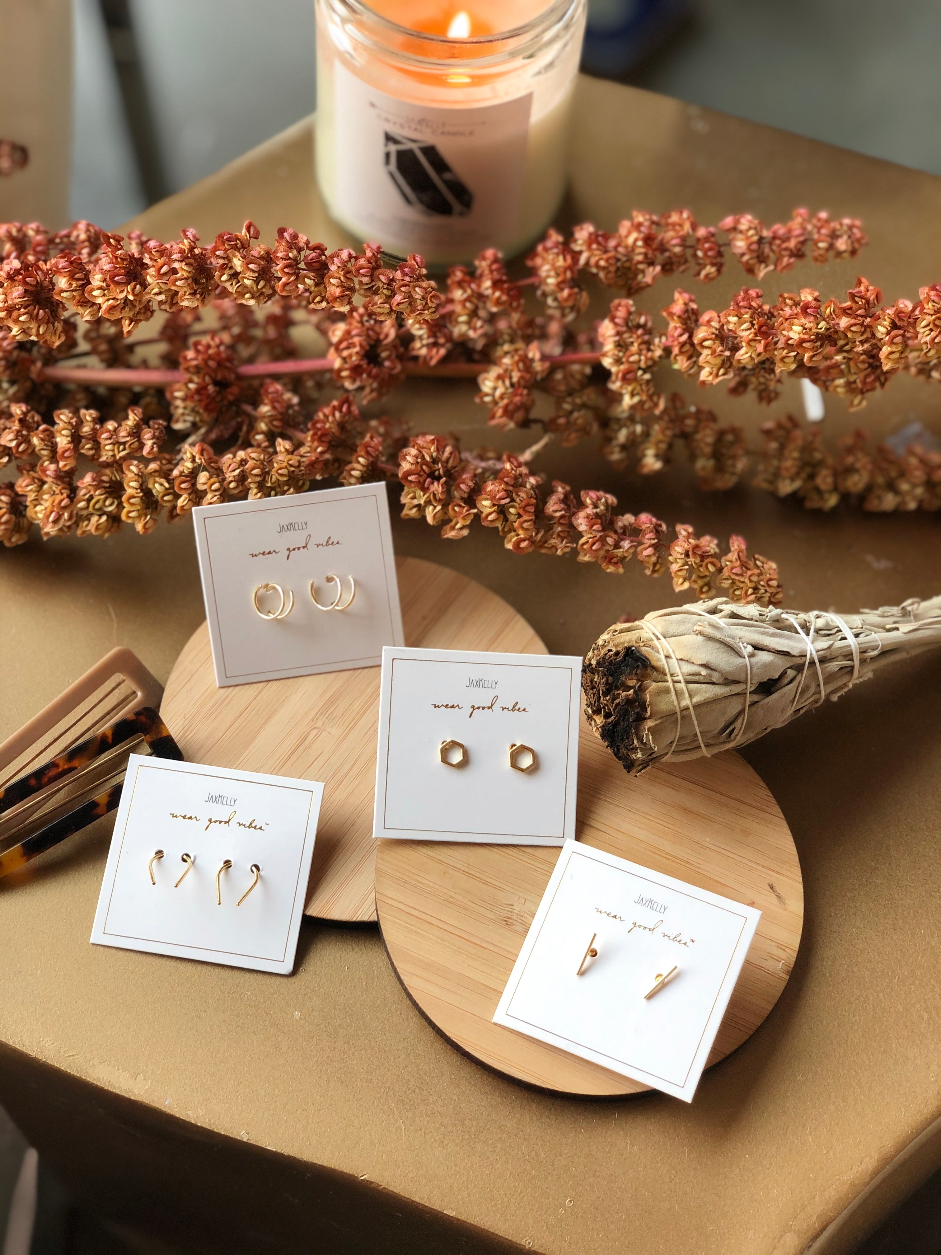 Gold earrings displayed on cards, arranged on round wooden trays, with dried floral decor, a bundle of sage, hair clips, and a lit candle in the background on a golden surface.