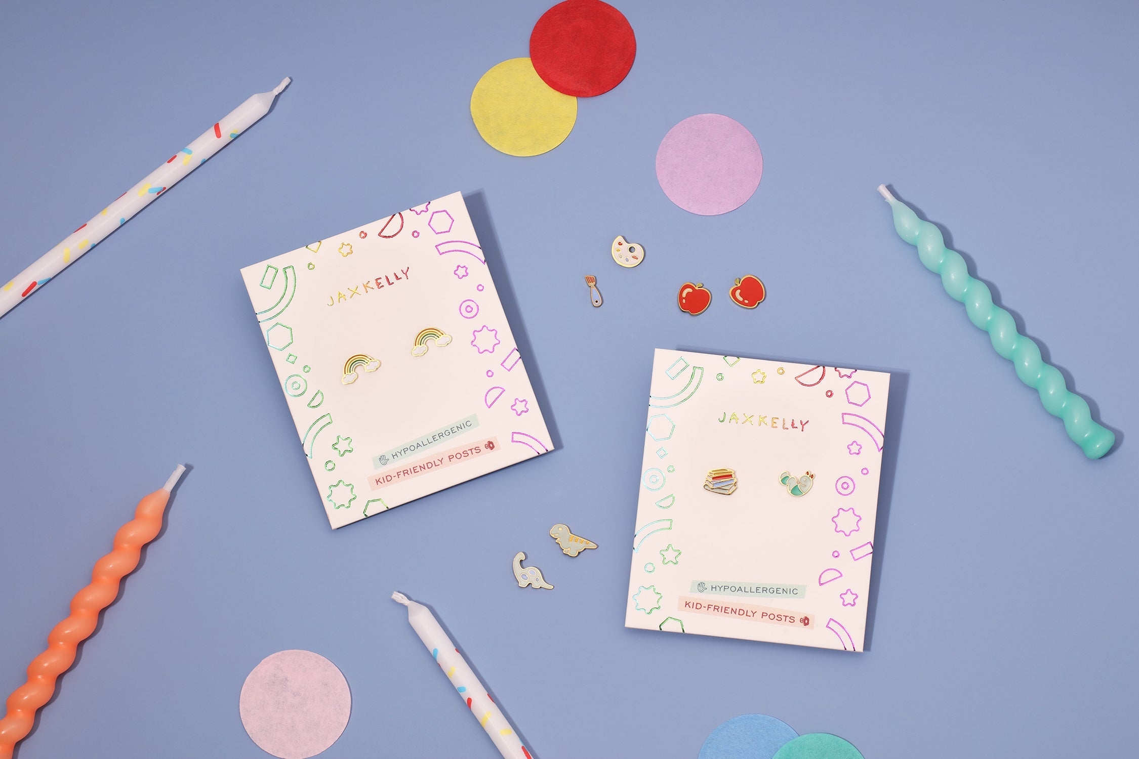 Two packs of kid-friendly earrings with colorful designs are placed on a light blue surface, surrounded by pastel twisted candles and bright paper confetti circles.