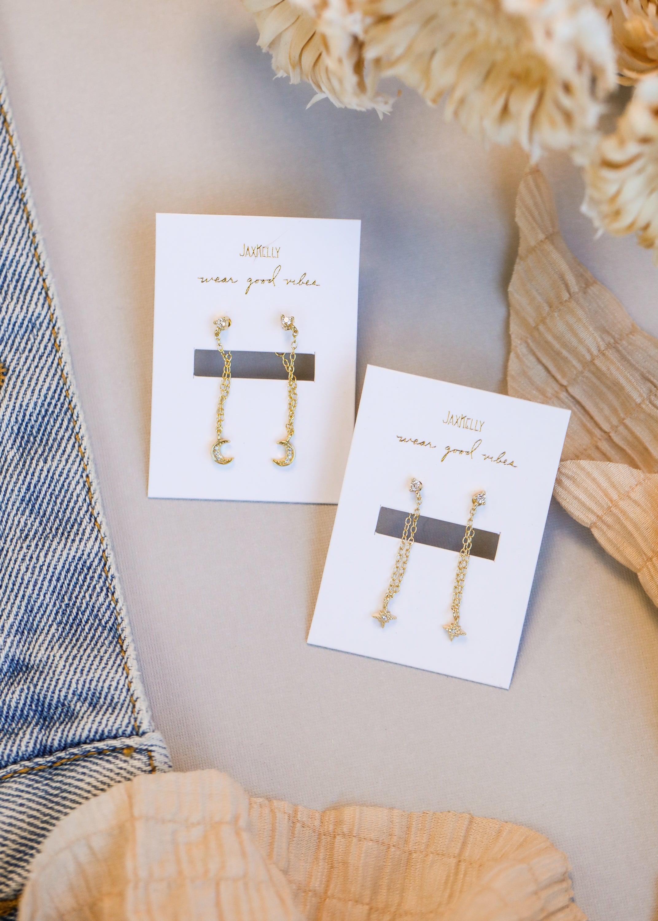 Two pairs of gold chain earrings are displayed on white cards; one pair features crescent moons, the other stars. The cards rest on a light surface, with fabric and denim partially visible.
