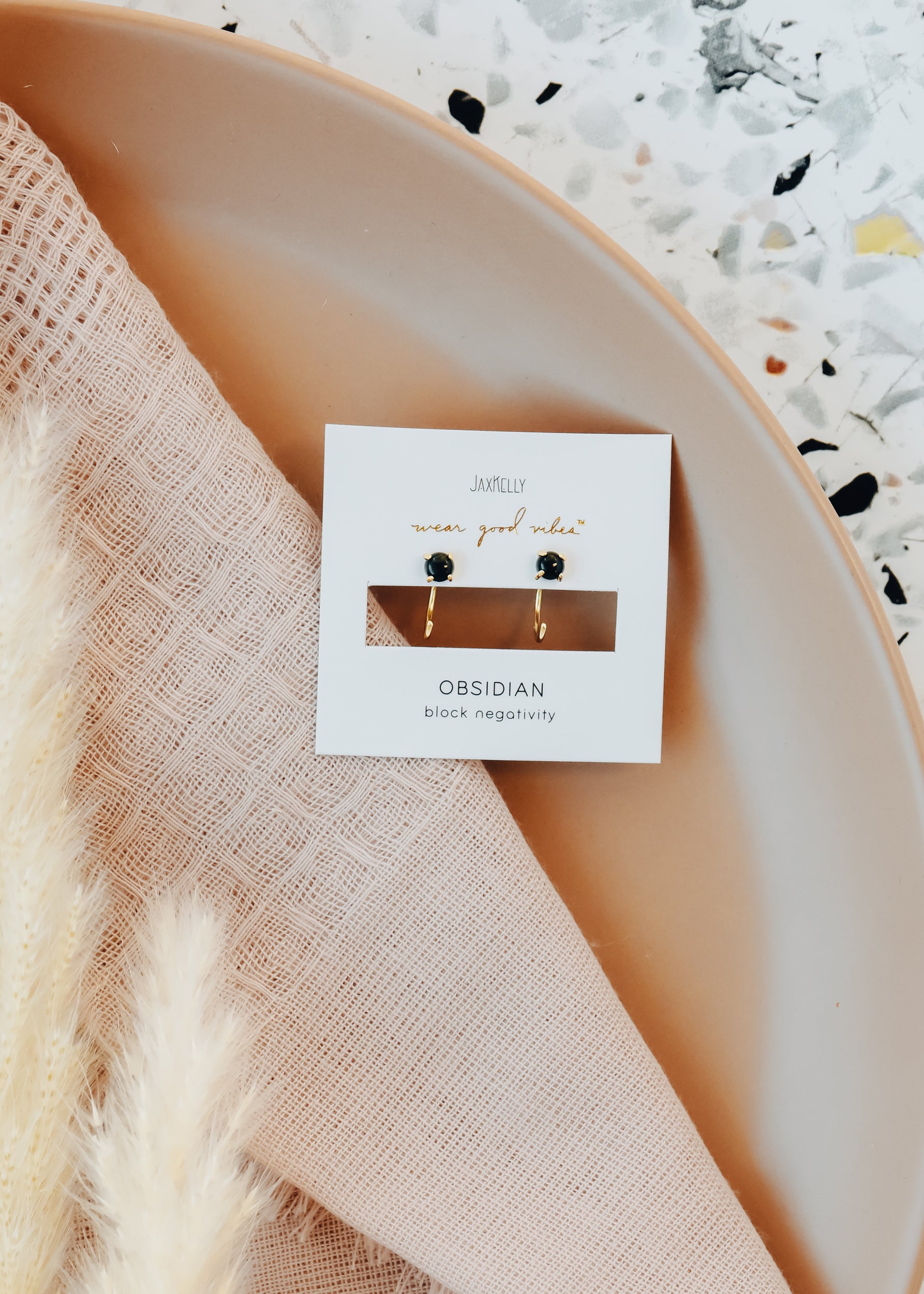 A pair of obsidian stud earrings on a white card, placed on a beige plate with a textured pink cloth and pampas grass nearby, on a speckled surface.