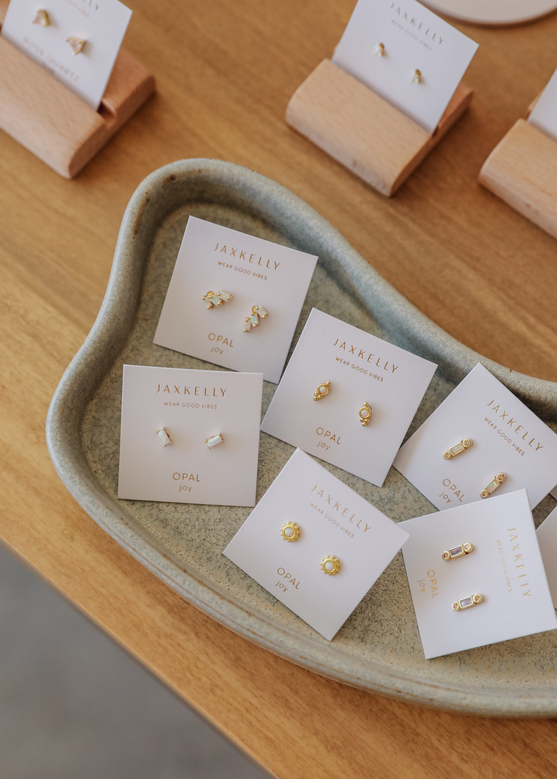 A ceramic tray holds several pairs of small gold and opal stud earrings on JaxKelly branded cards, displayed on a wooden surface. More earrings are in the background.