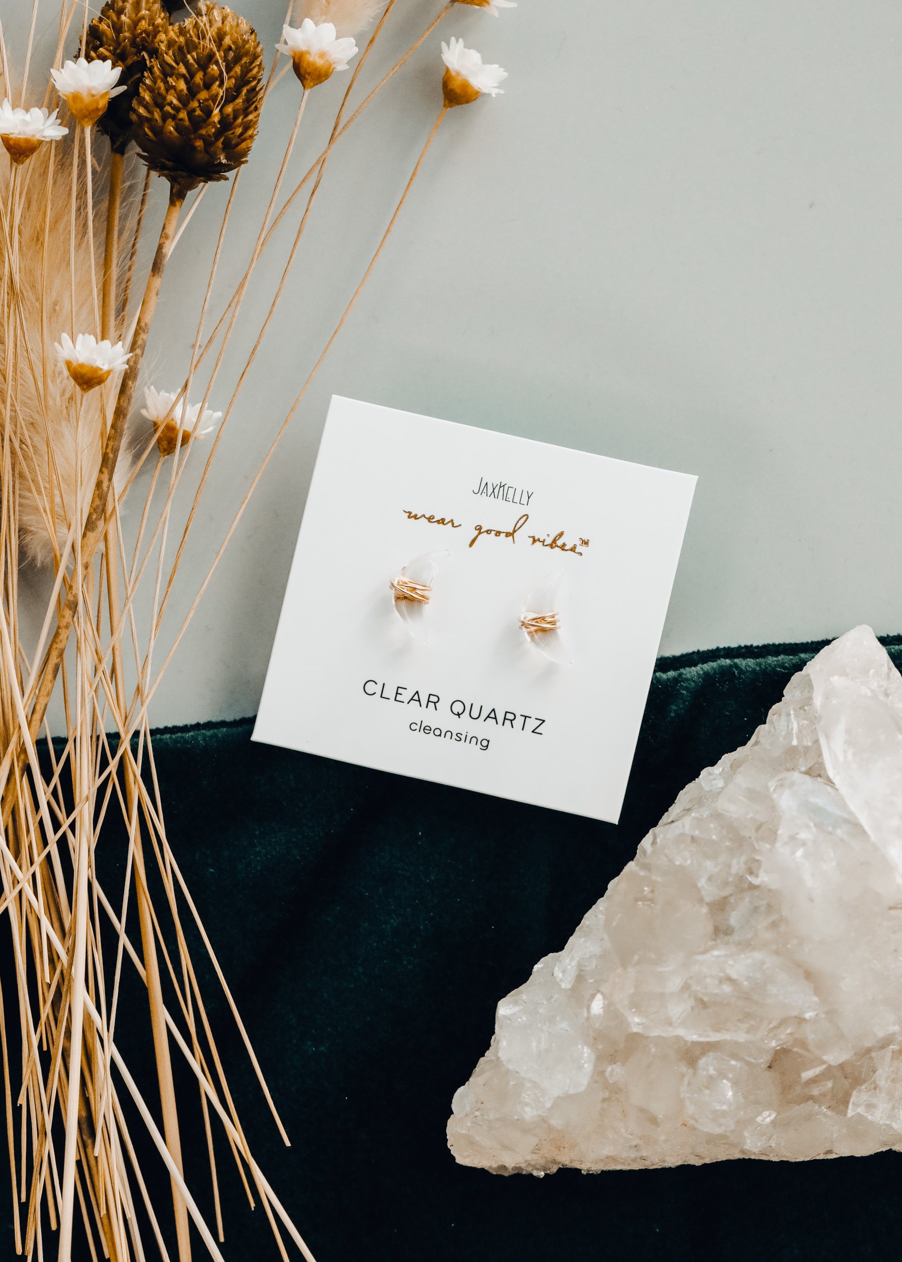 A pair of clear quartz stud earrings on a card with gold text, next to a large quartz crystal and dried wildflowers on a dark velvet cloth.