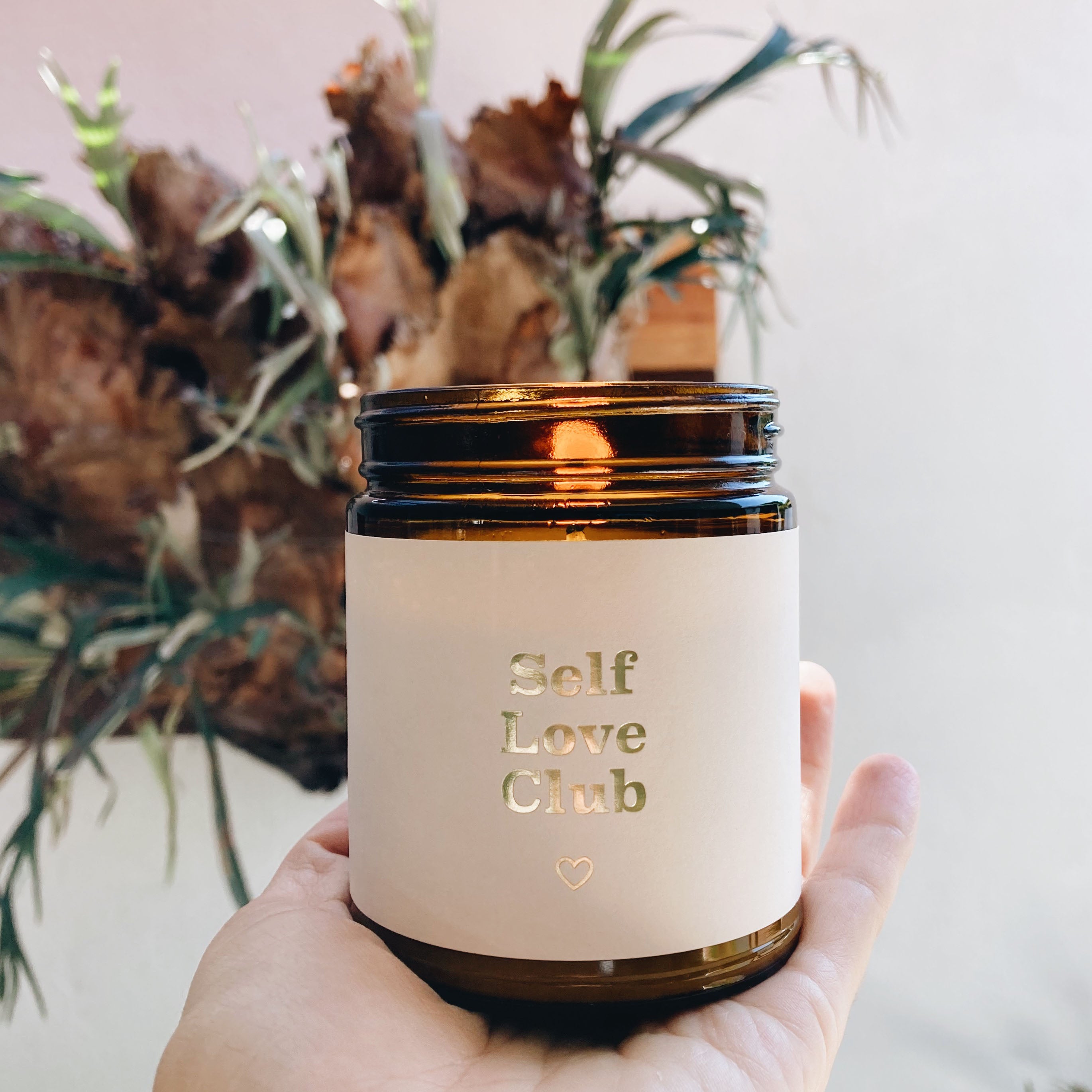 A hand holds a brown glass jar candle with a label that reads Self Love Club in gold letters, with a small heart below. Blurred leafy plant in the background.