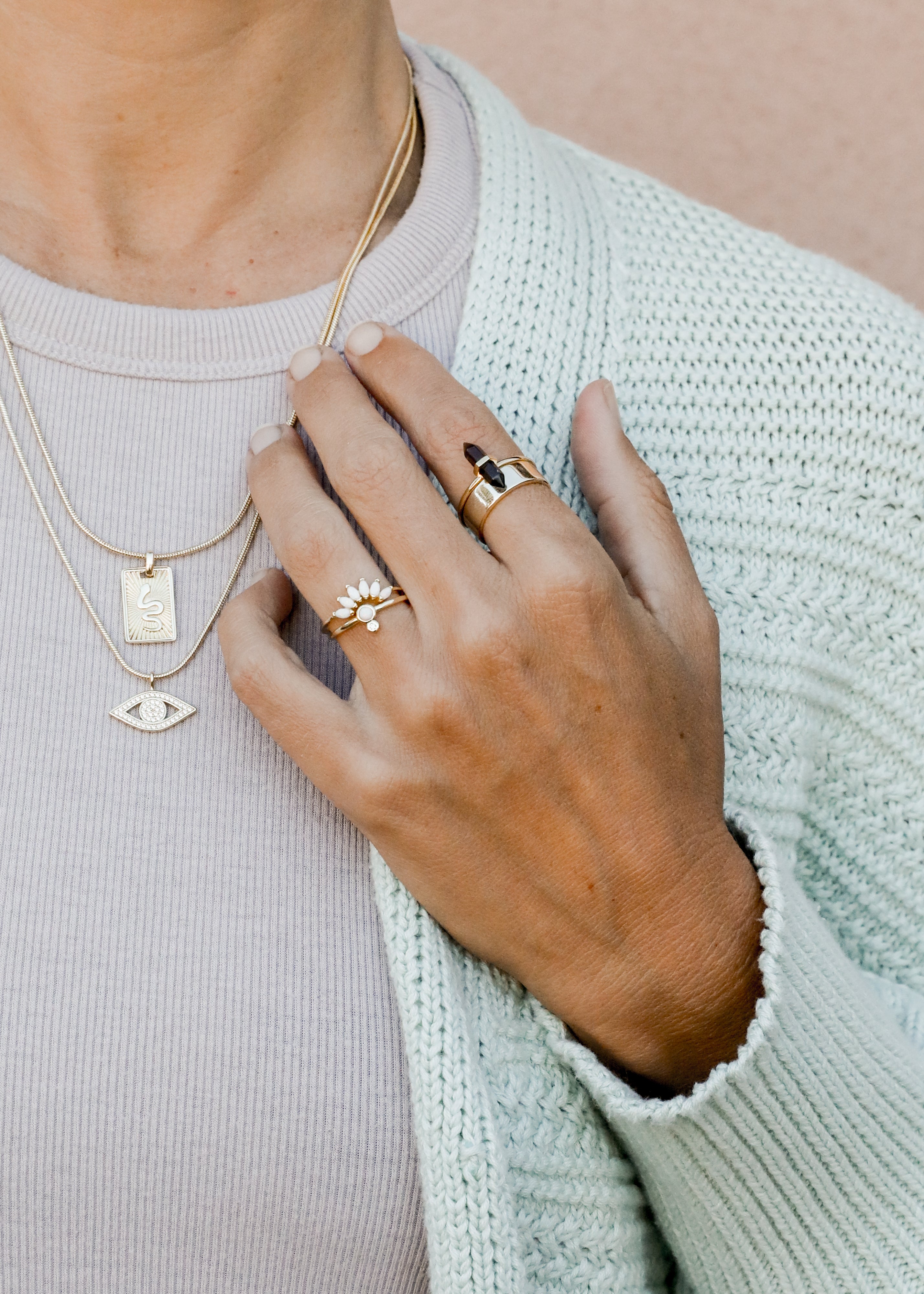 A person wearing layered gold necklaces and multiple gold rings on their hand, dressed in a light knit cardigan and a ribbed top, with their hand gently touching their chest.