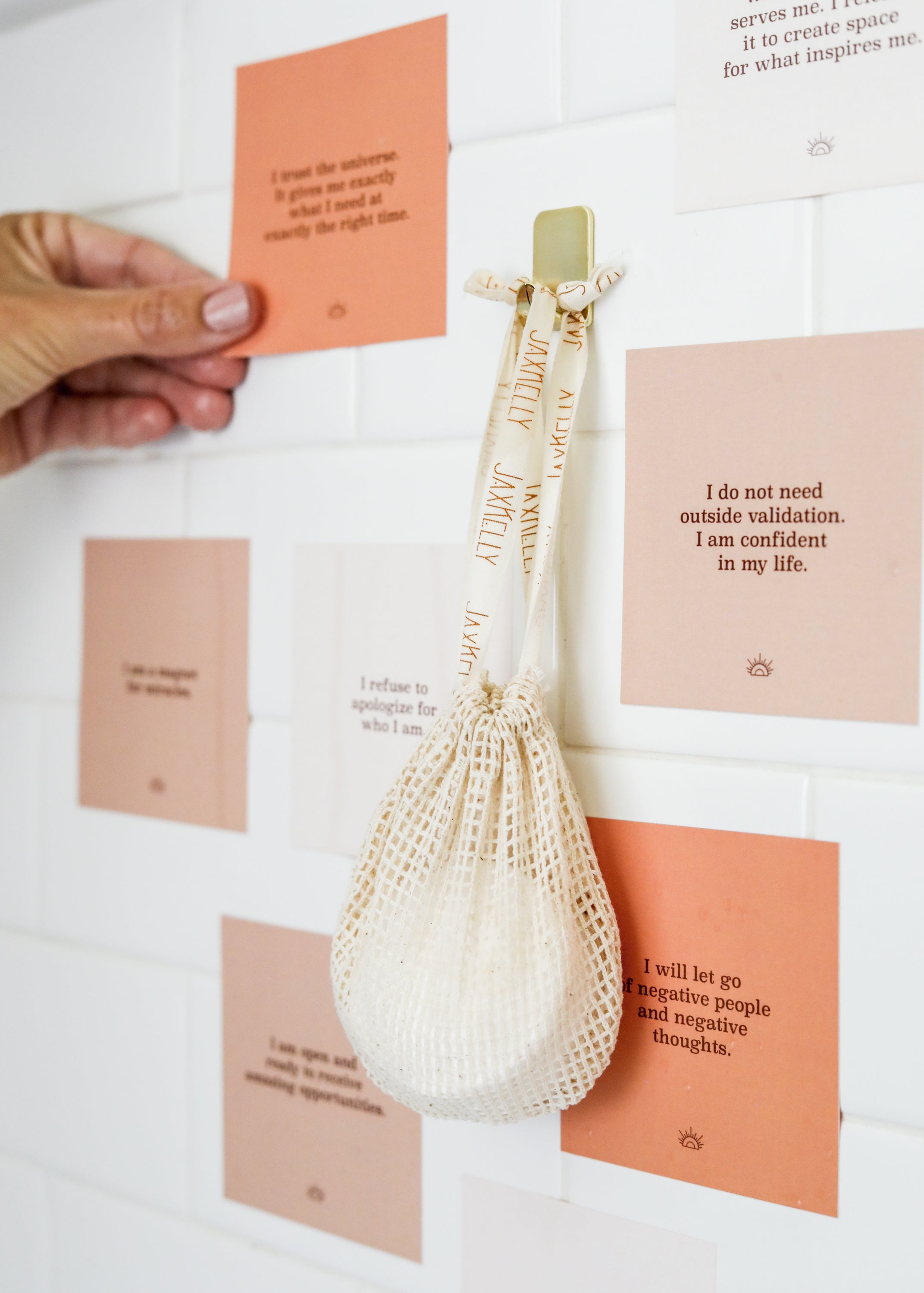 A hand sticks an inspirational quote card on a white tiled wall decorated with other motivational cards. A mesh bag with a round object hangs from a hook among the cards.