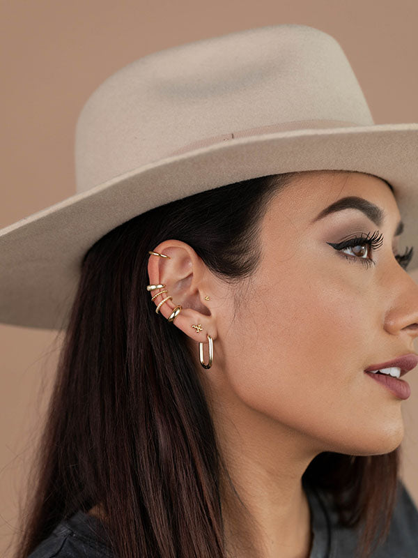 Woman in a beige hat with long dark hair, wearing multiple gold earrings and ear cuffs, looking to the right. She has winged eyeliner and a neutral lipstick shade.