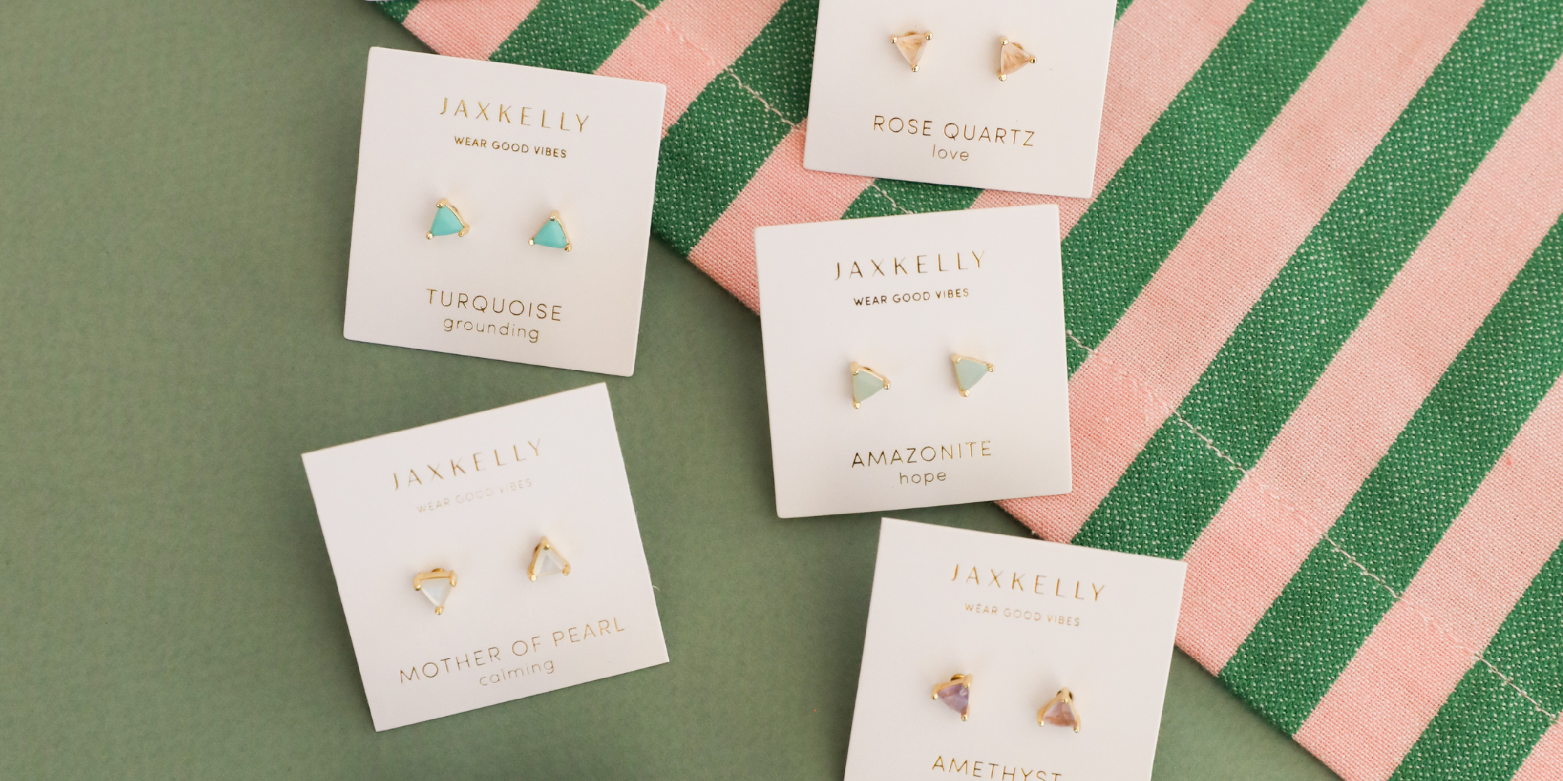 Five pairs of gemstone stud earrings on JaxKelly cards are arranged on a green and pink striped fabric background. Each card lists the stone name and a positive word like “hope” or “love.”.