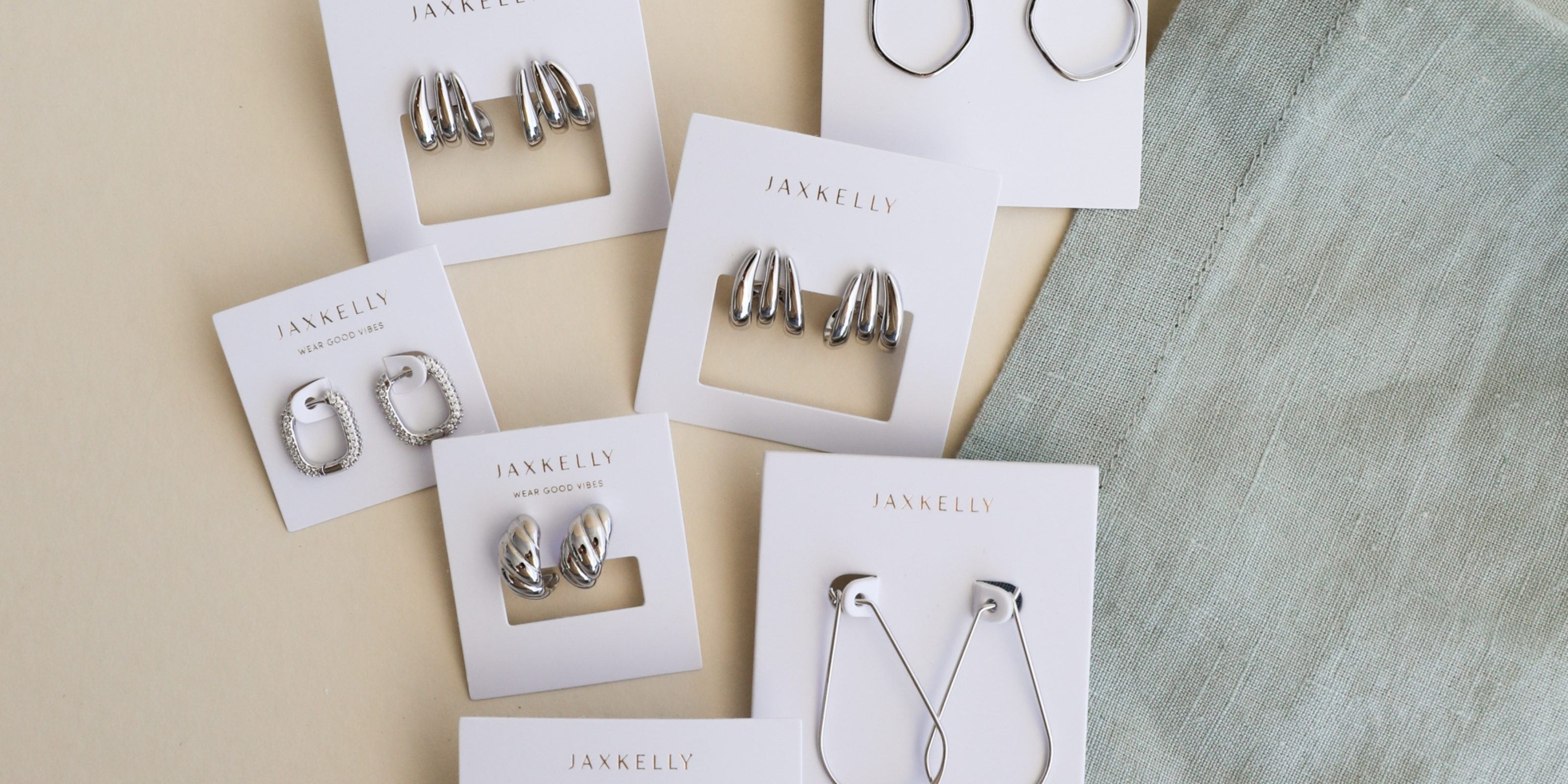 A flat lay of six pairs of silver earrings on white JaxKelly cards, arranged on a cream surface next to a light blue textured fabric.
