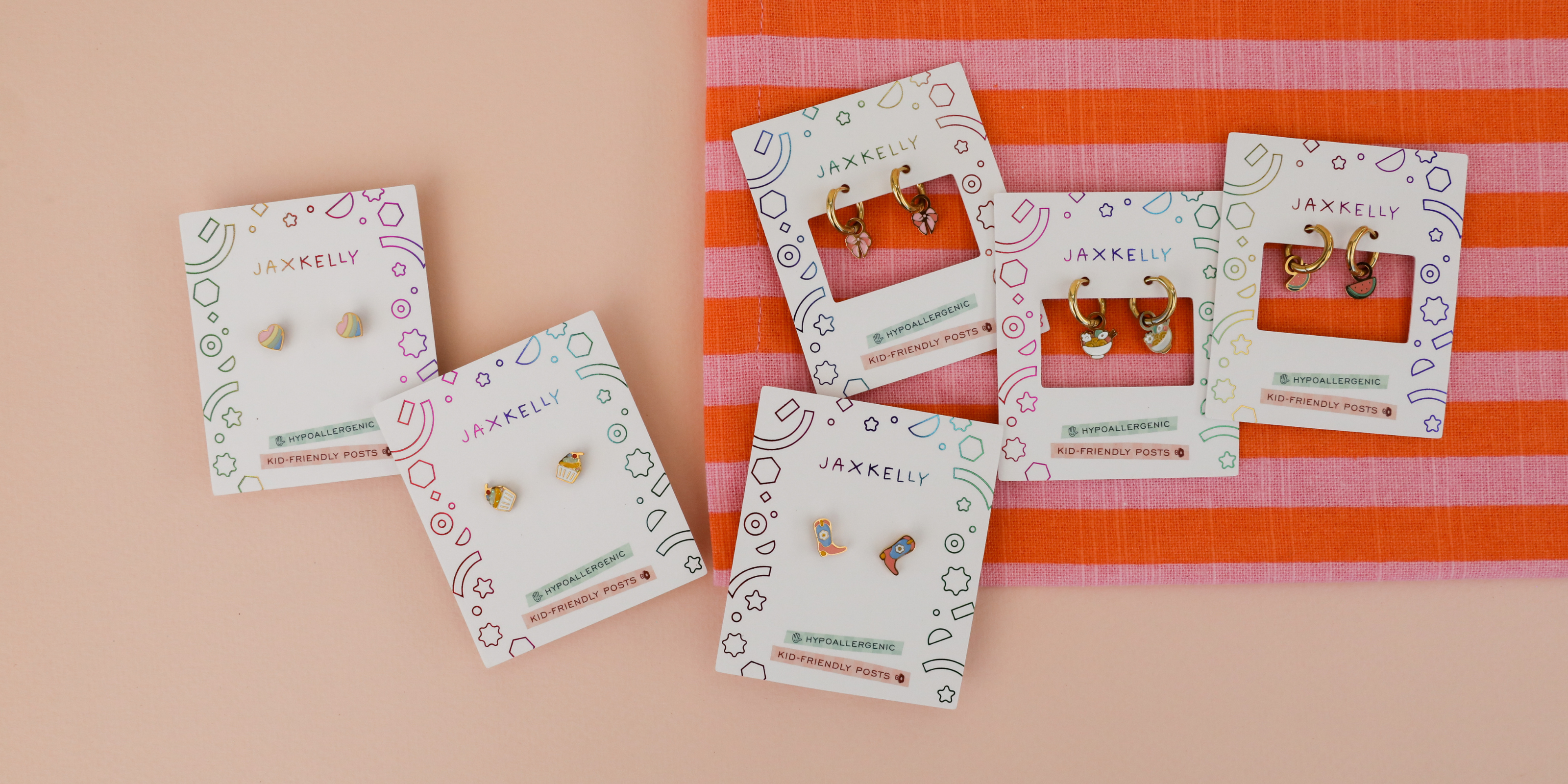 Six pairs of colorful earrings on JaxKelly display cards are arranged on a peach background with a pink and orange striped cloth in the top right corner.