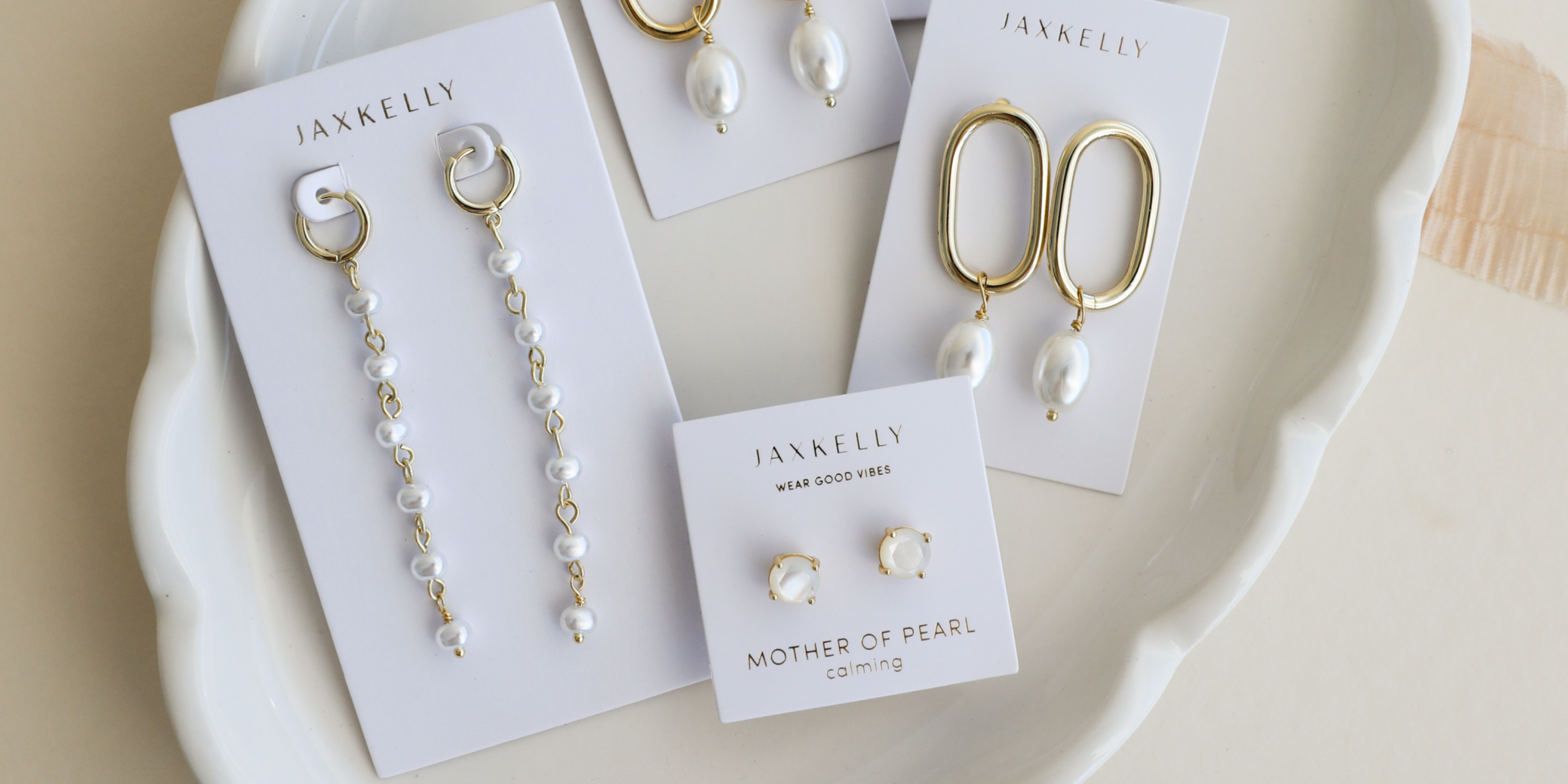 Four sets of JaxKelly earrings, including dangling pearl designs and gold hoop styles, displayed on white cards in a white dish. One card reads Mother of Pearl with small pearl stud earrings.