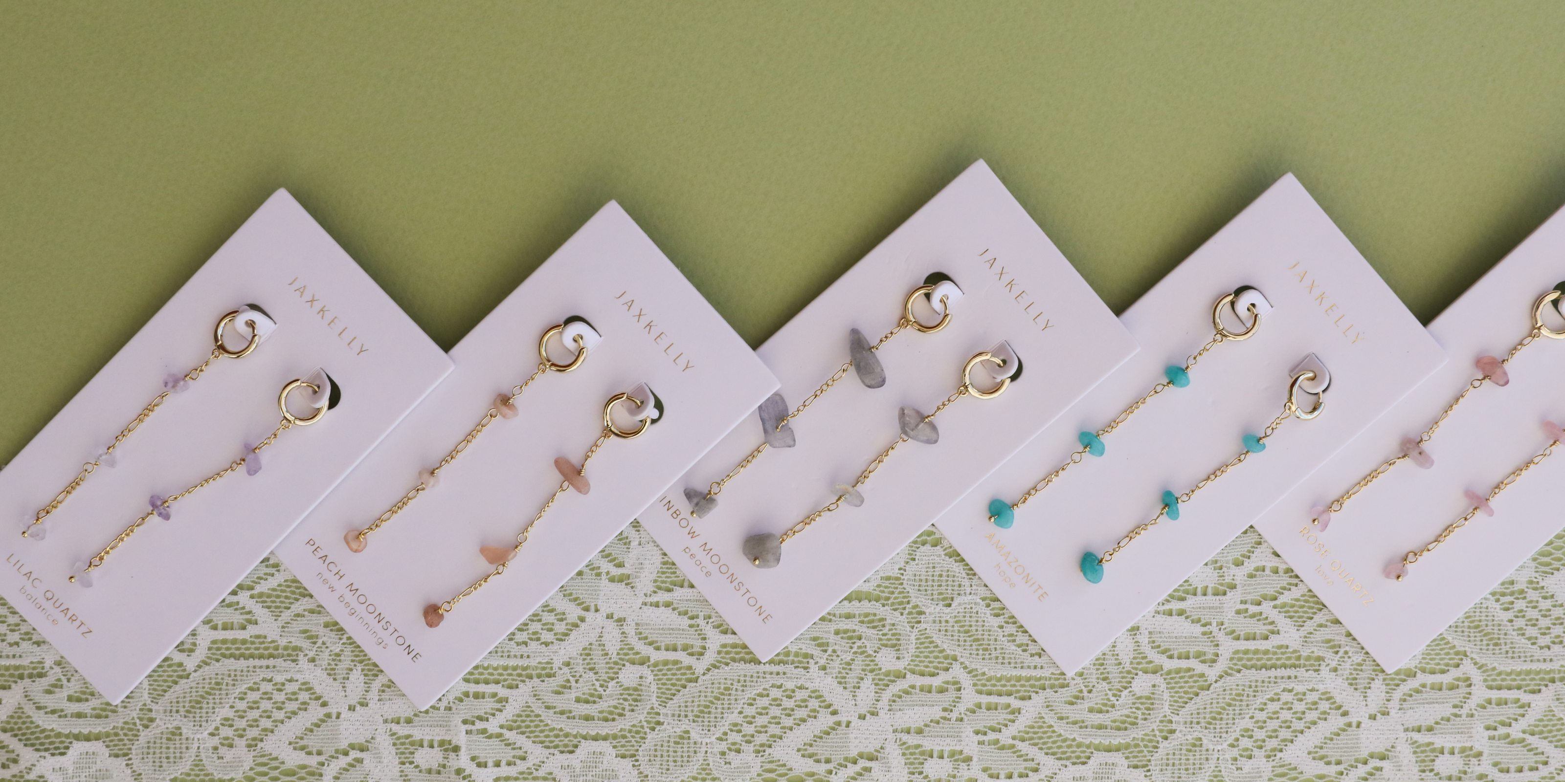 Five pairs of dangly earrings with various pastel-colored stones are displayed on white cards against a green background and lace fabric. The earrings are arranged in a neat, angled row.