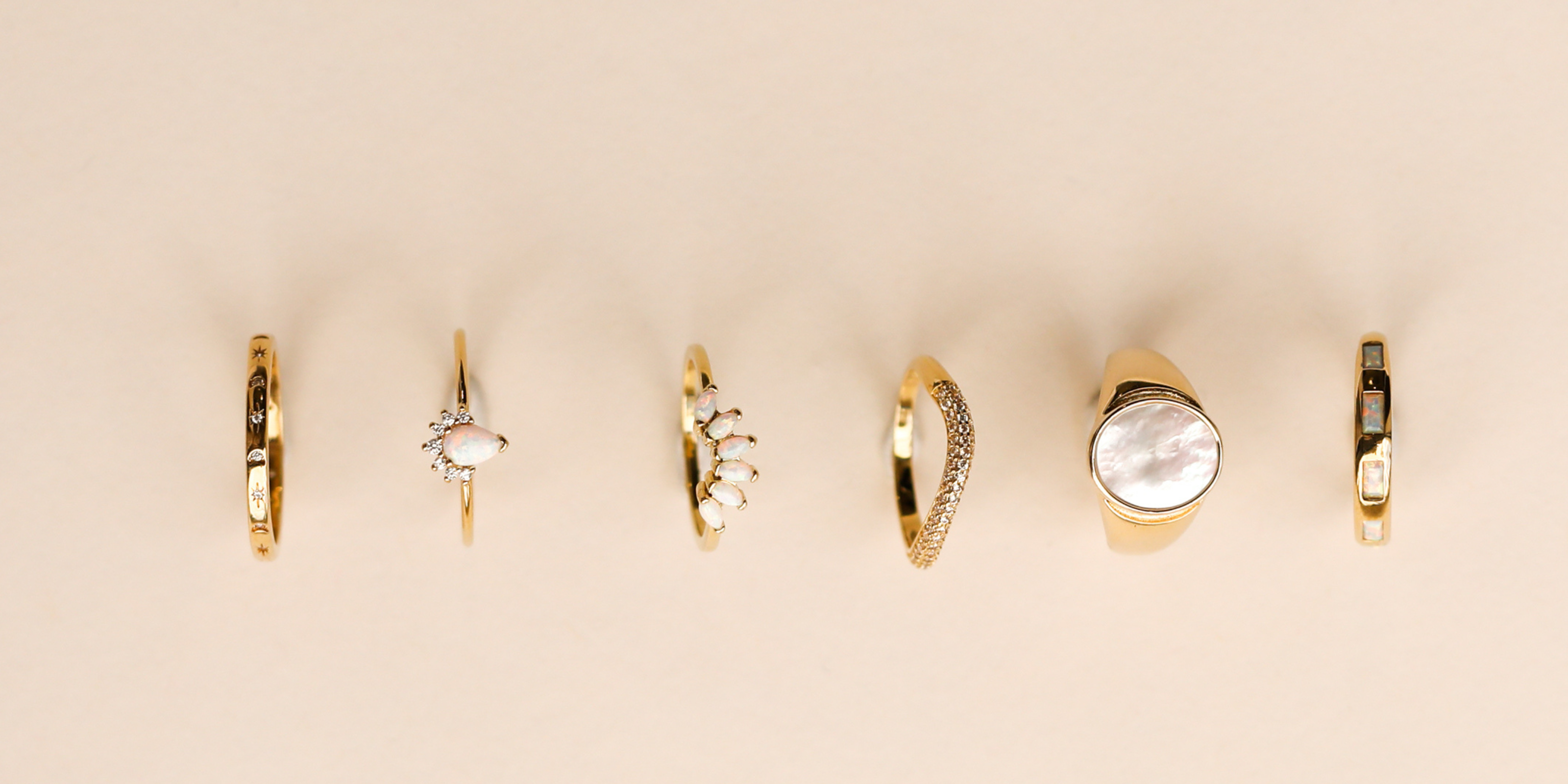 Six gold rings with various gemstone and pearl designs are arranged in a row on a light beige background.