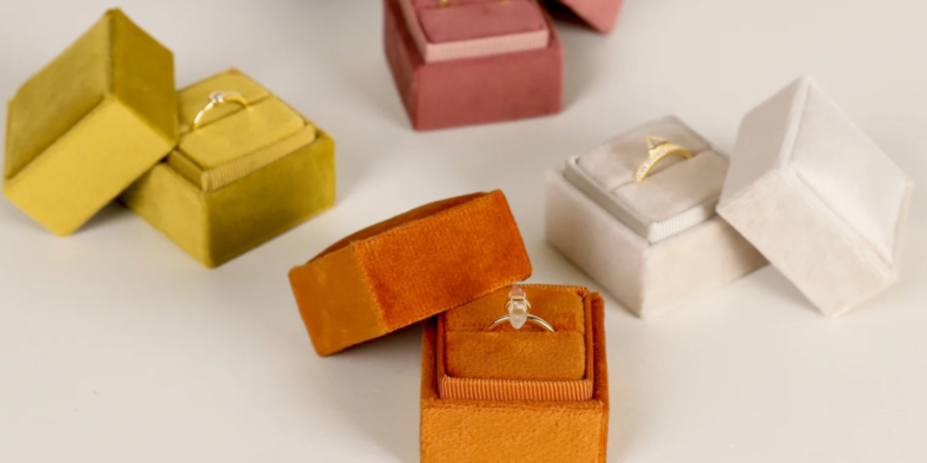 Several colorful velvet ring boxes, each holding a different ring, are arranged on a light surface. The boxes are in shades of orange, mustard yellow, pink, and off-white.