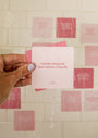 A hand with light-purple nail polish holds a pink Self-Love Shower Affirmations™ waterproof card reading "I am becoming the best version of myself." Water droplets and more pink reusable shower cards appear on white tiled walls in the background.