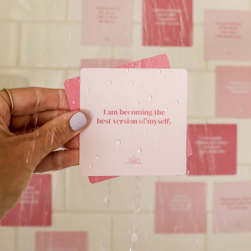 A hand with light-purple nail polish holds a pink Self-Love Shower Affirmations™ waterproof card reading "I am becoming the best version of myself." Water droplets and more pink reusable shower cards appear on white tiled walls in the background.