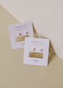A pair of White Opal Huggie earrings is displayed on a white JaxKelly card atop a beige and off-white surface.