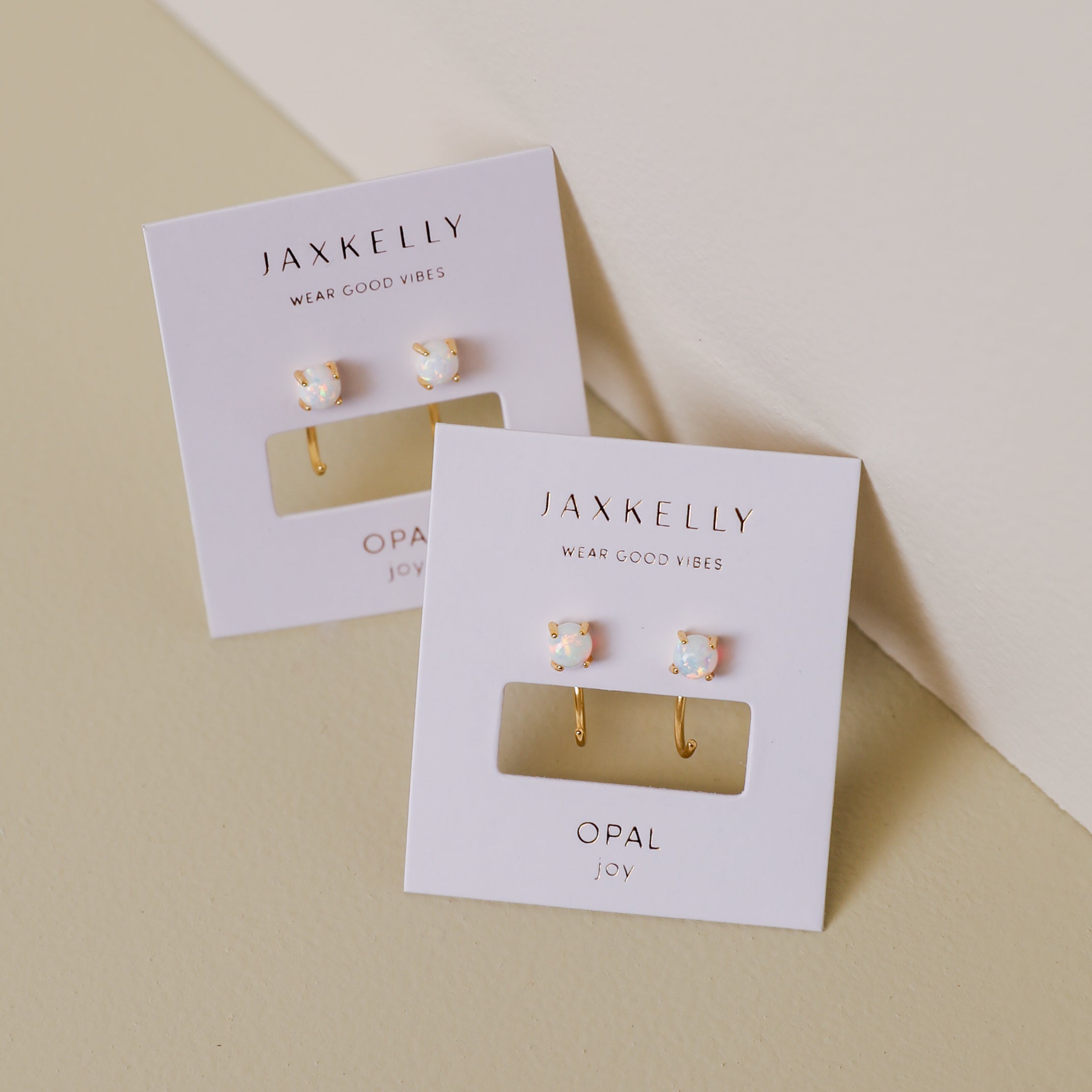 A pair of White Opal Huggie earrings is displayed on a white JaxKelly card atop a beige and off-white surface.