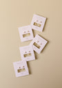 Five pairs of White Opal Huggie earrings are displayed on cream-colored JaxKelly cards, arranged diagonally against a beige background—ideal for those who love gold plated huggies or white opal jewelry.
