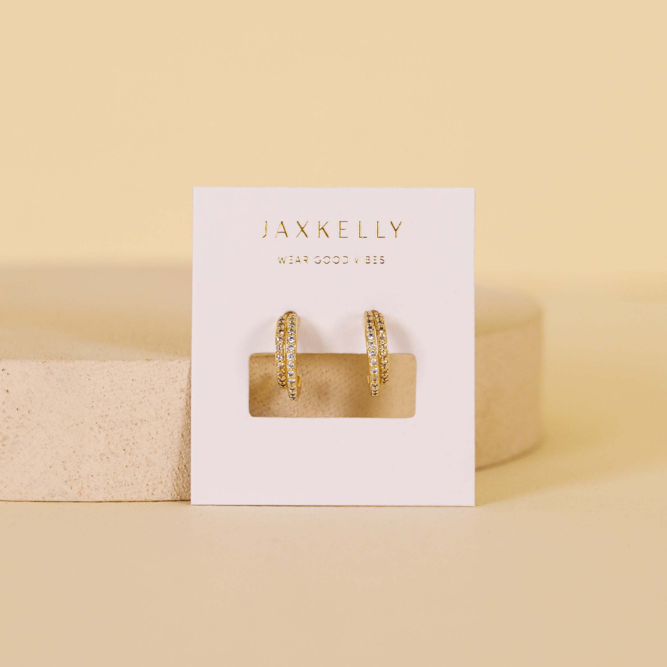 The Double Champagne Hoop - Earrings feature textured double offset hoops with 18K gold plating, displayed on a white JaxKelly card atop a round platform set against a soft beige background.