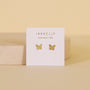 A pair of Butterfly gold stud earrings with sterling silver posts, displayed on a white JaxKelly card reading "Wear Good Vibes," set against a soft beige background.