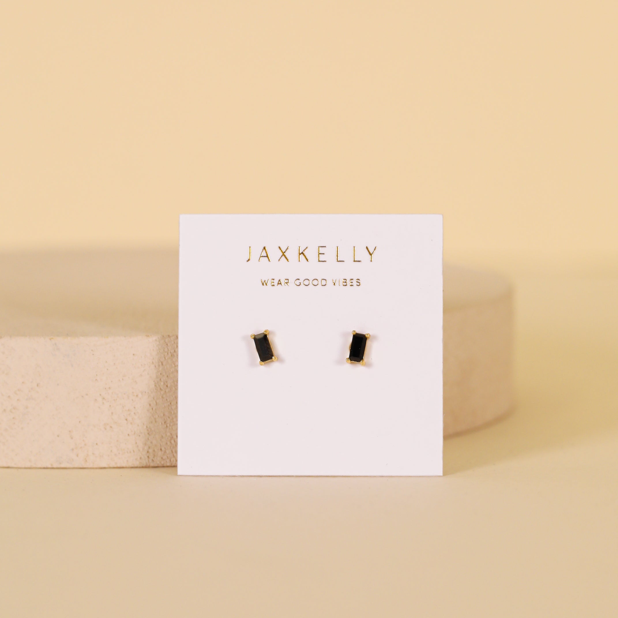 The Black Baguette earrings are rectangular black baguette studs with 18k gold plating and sterling silver posts, shown on a white JaxKelly card against a soft beige background.