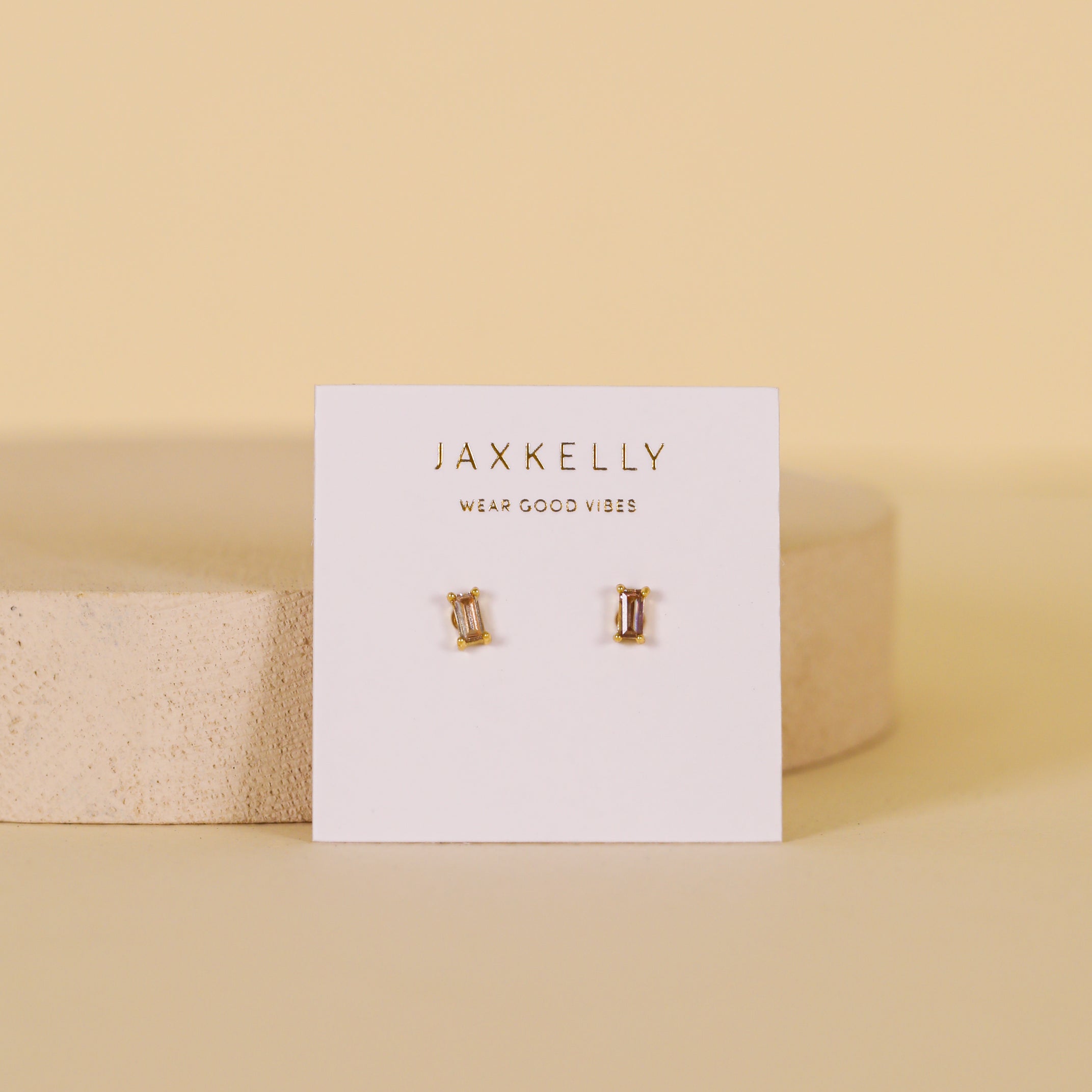 A pair of small gold plated Coffee Baguette earrings with sterling silver posts, displayed on a white JaxKelly card featuring the slogan "Wear Good Vibes," set against a soft beige background.