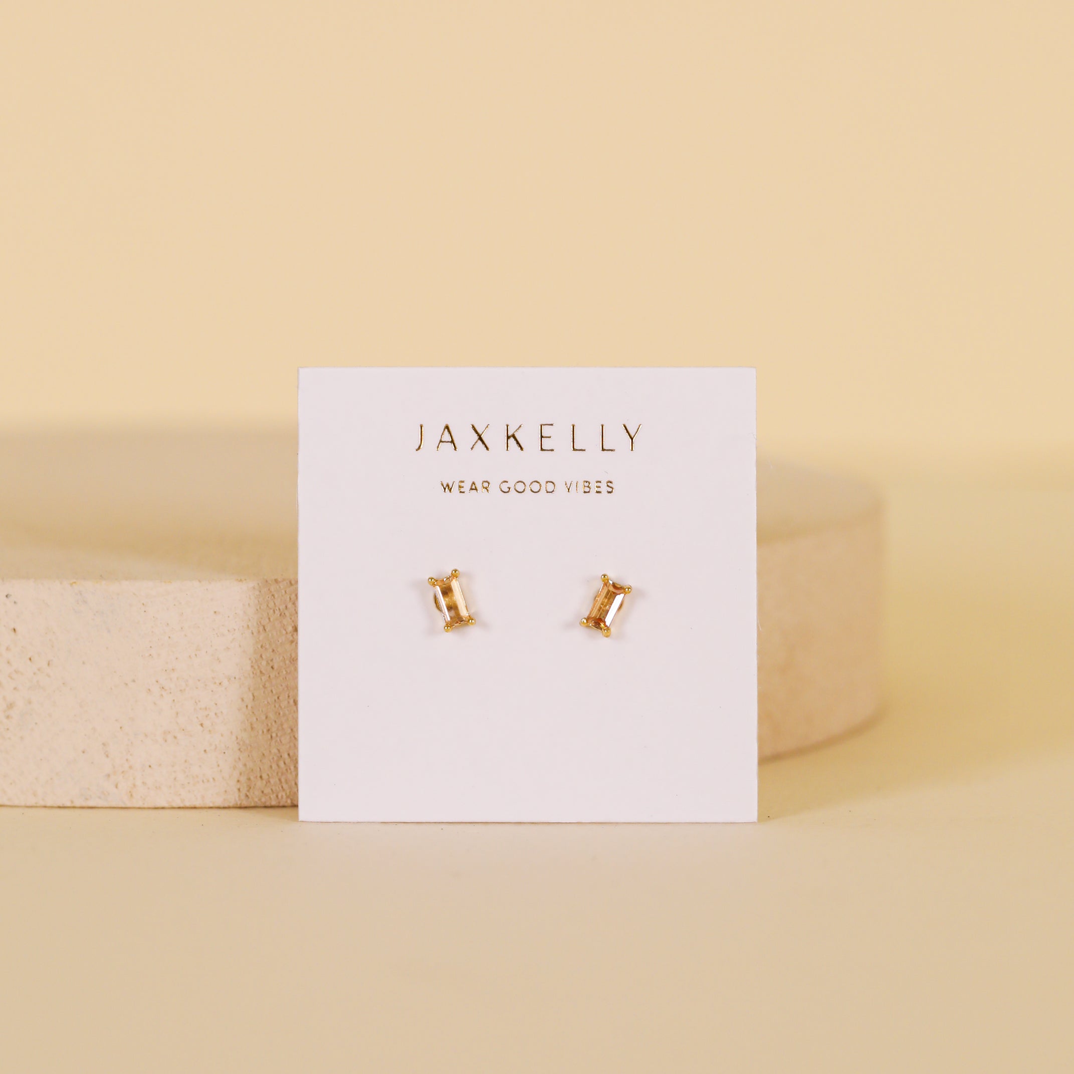 A pair of Champagne Baguette gold plated stud earrings with sterling silver posts are displayed on a white JaxKelly Wear Good Vibes card, set on a soft beige background with a round platform.