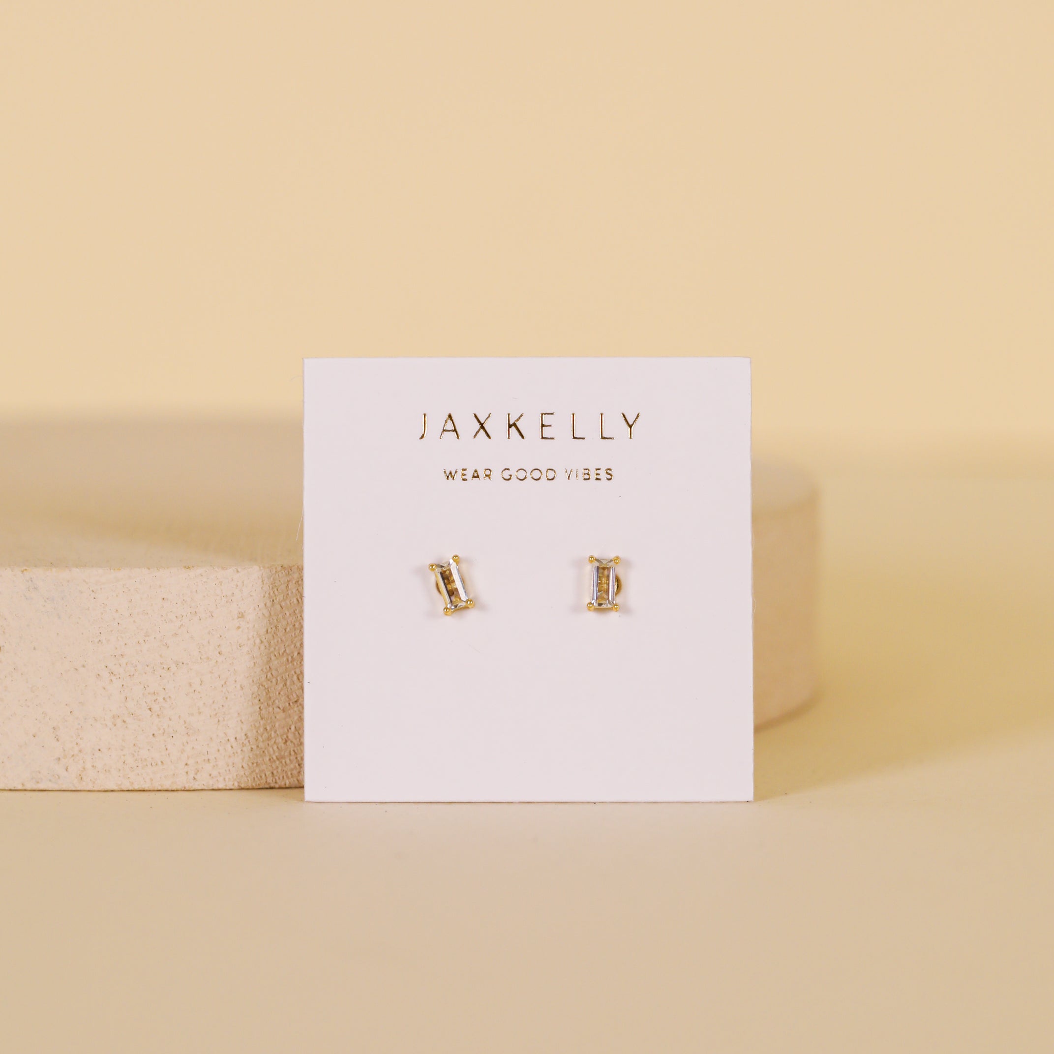 A pair of Clear Baguette stud earrings with 18k gold plating and sterling silver posts, displayed on a white JaxKelly card reading WEAR GOOD VIBES, set against a soft beige background.