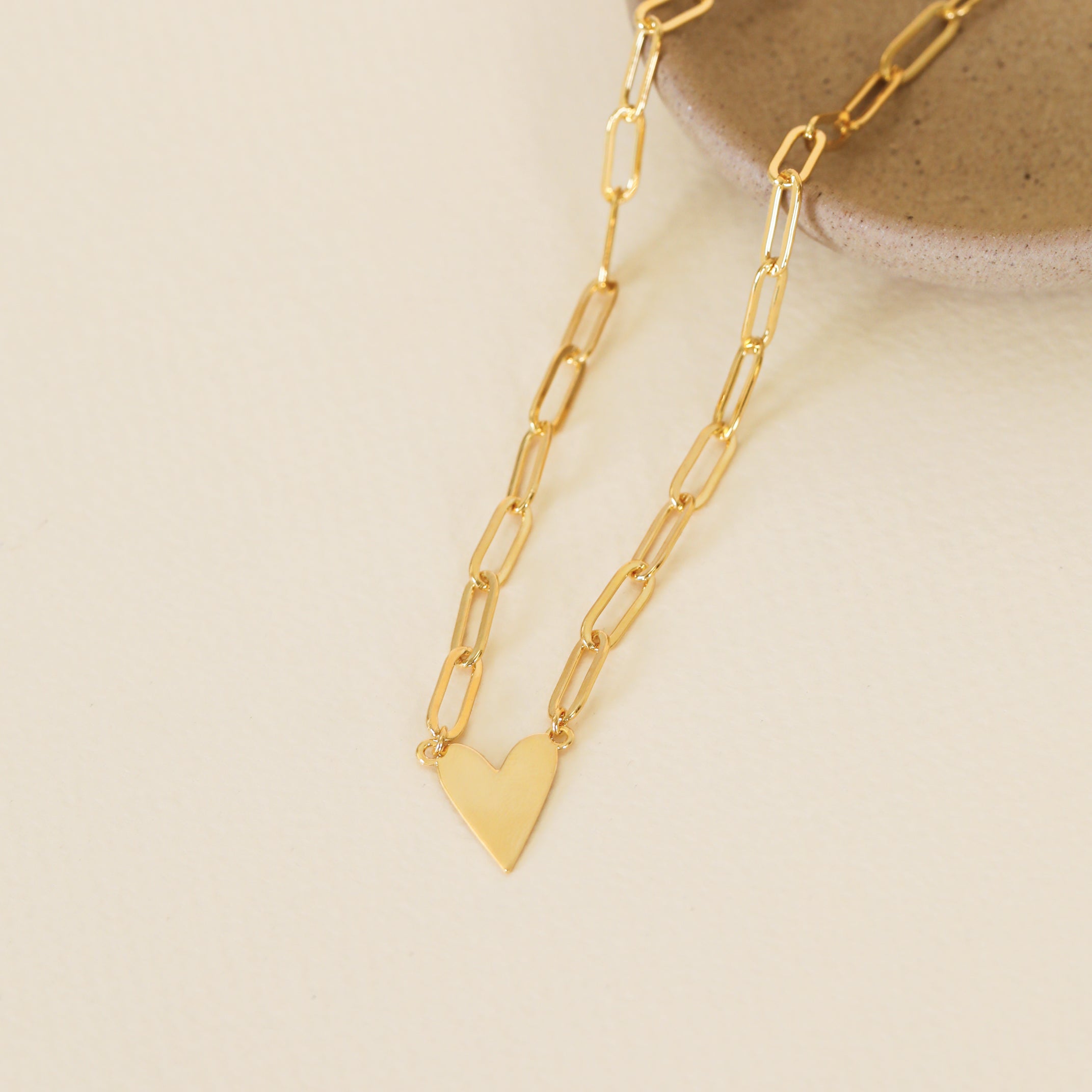 The Gold Heart Pendant Link Chain Necklace - 18k Gold Plated features elongated links and a small heart charm, displayed on a round beige ceramic dish against a light cream background.